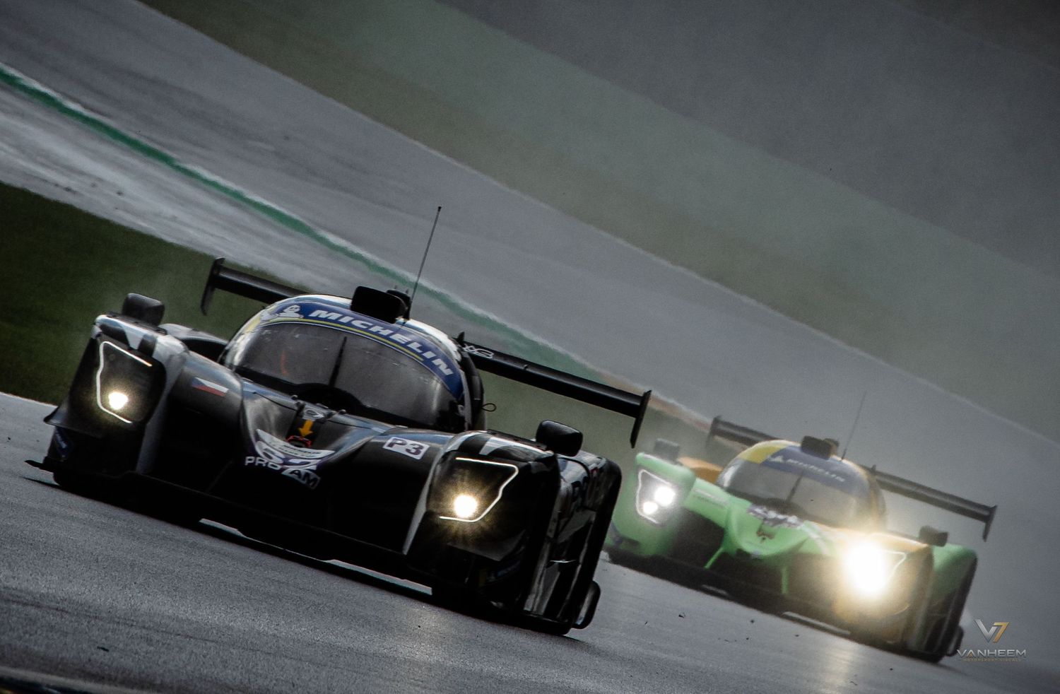 Two LMP racing cars compete on a wet track at night, headlights blazing in dramatic motorsport action.