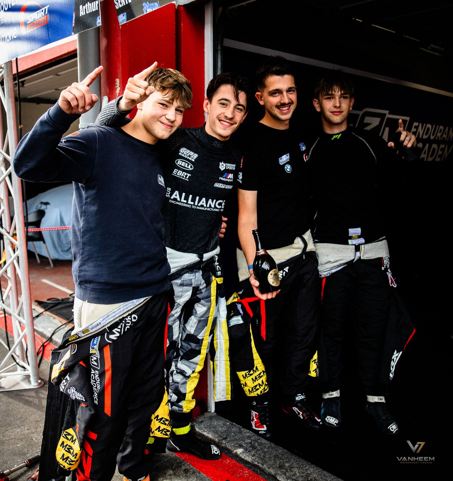 Four young racing drivers in karting suits pose together smiling at a motorsport event paddock area.