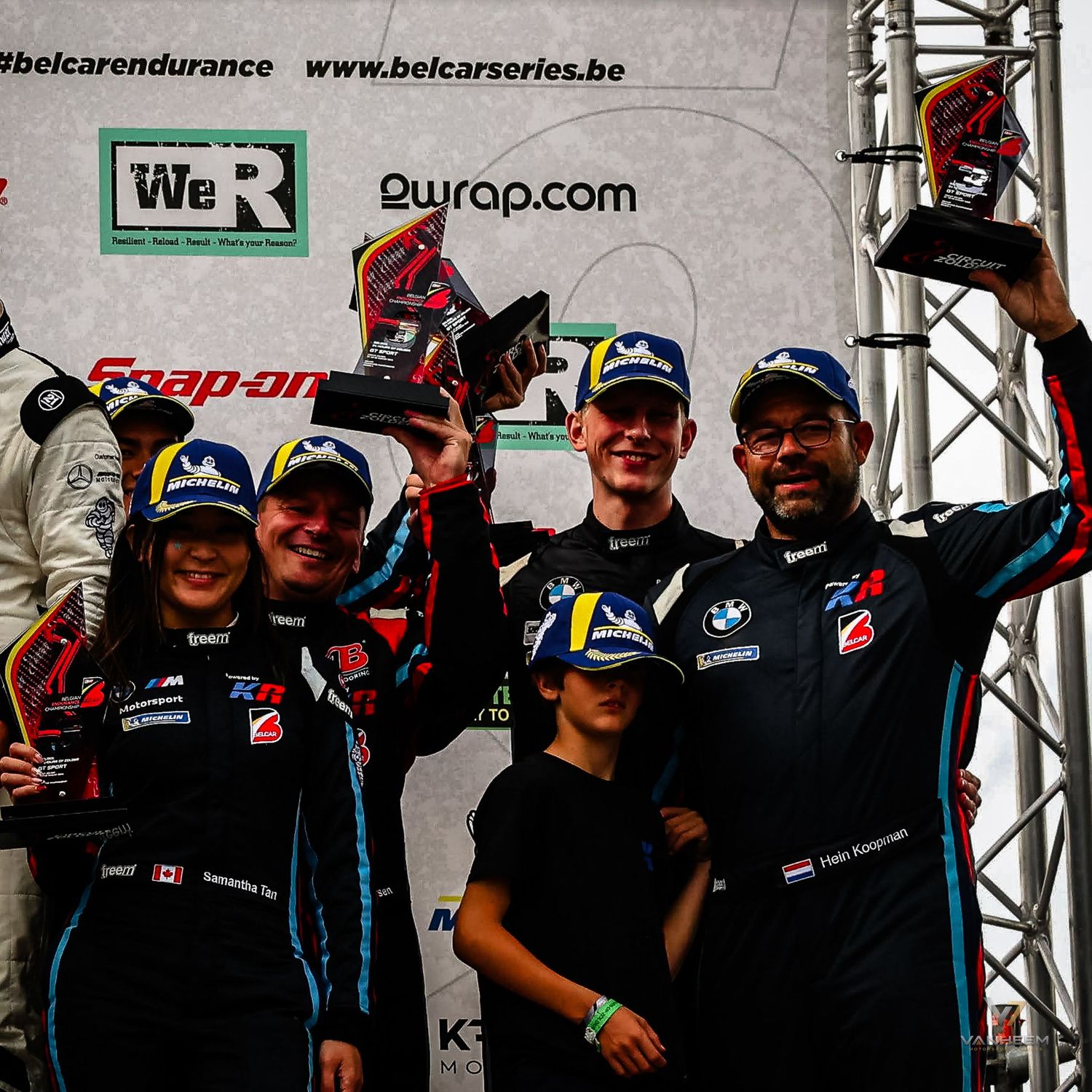 Racing team celebrates victory on podium holding trophies at Belcar Endurance Series event with sponsors visible.
