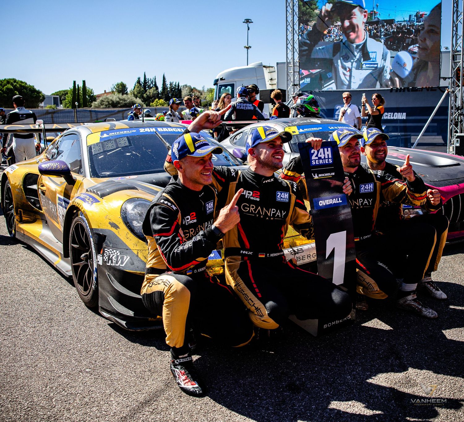 Racing team celebrates victory beside a yellow and black Porsche GT3 race car marked with number 1 at a motorsport event.