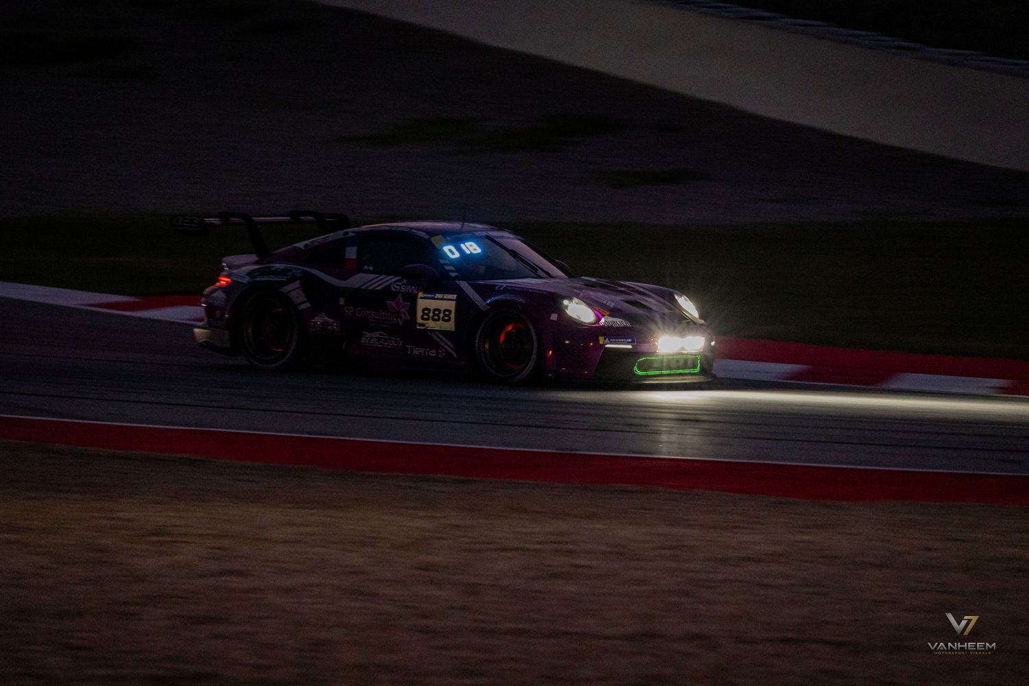 A dark Porsche GT3 race car speeds around a circuit track at night under bright headlights.