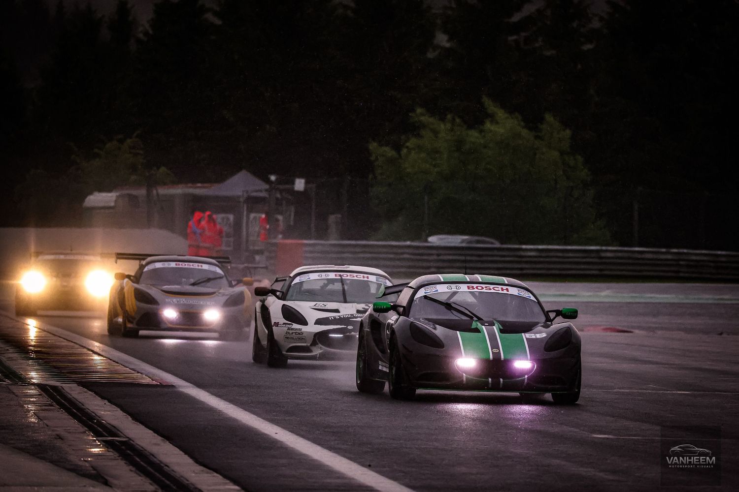 Racing cars speed on a wet track at dusk, with headlights glowing and a dark green sports car leading the pack.