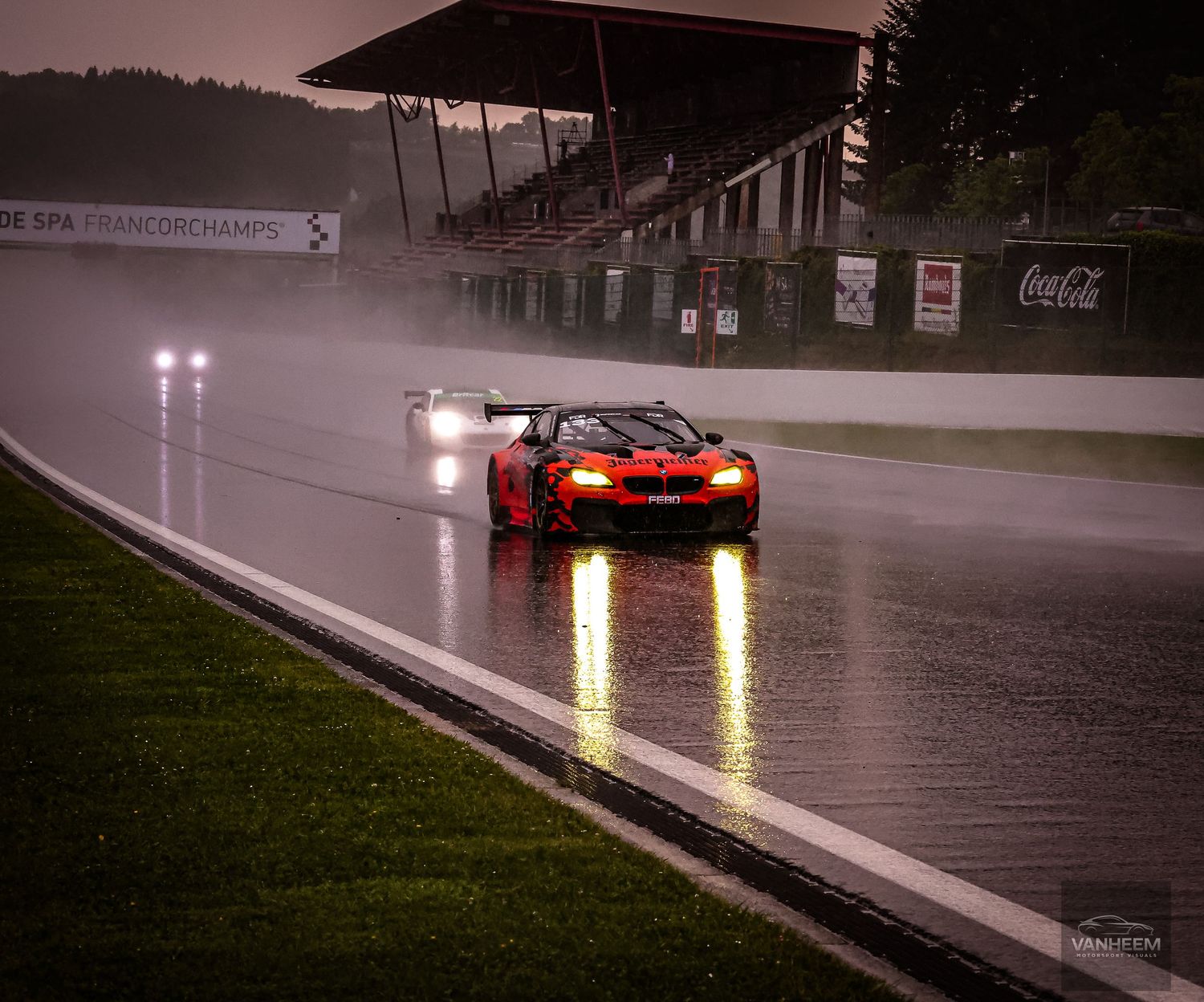 Red GT race car speeding on wet Spa-Francorchamps track in heavy rain during endurance race.