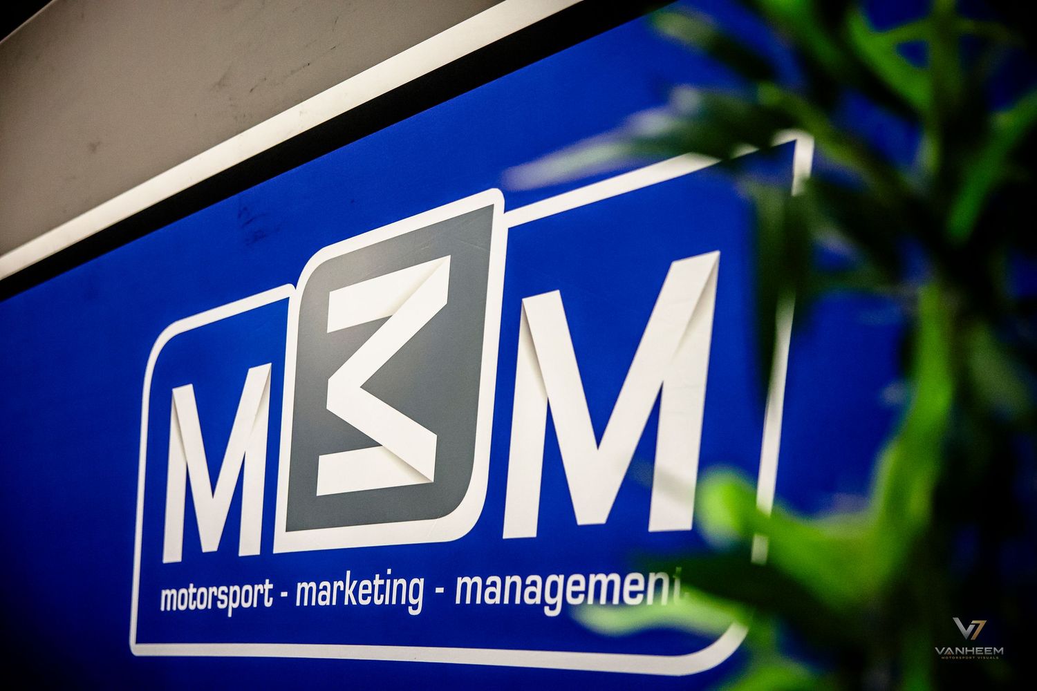 Blue M3M Motorsport Marketing Management logo sign displayed outdoors with green foliage in foreground.
