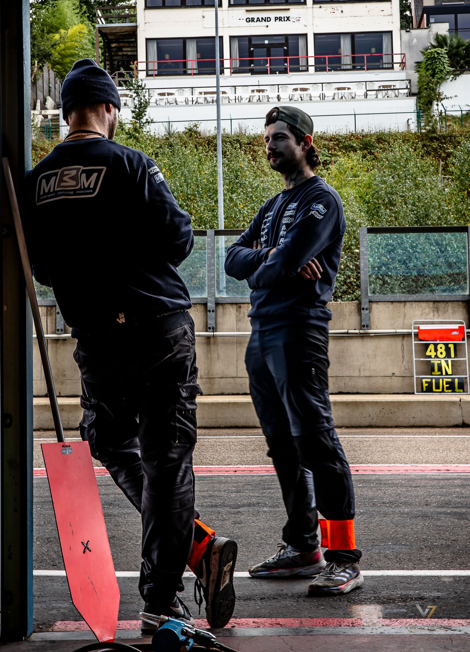 Two mechanics in dark uniforms discuss strategy in a Formula 1 pit lane, with a red lollipop sign and fuel display visible.