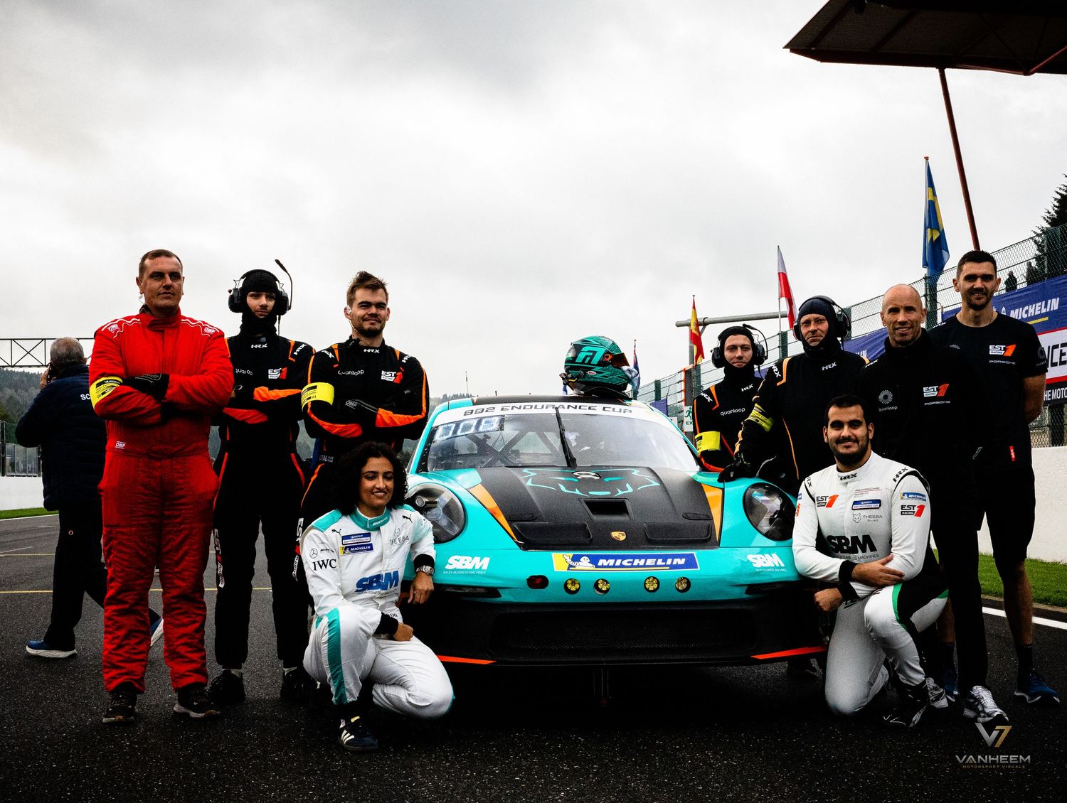 Racing team poses with a Porsche GT car on a wet track, wearing motorsport gear and race suits.