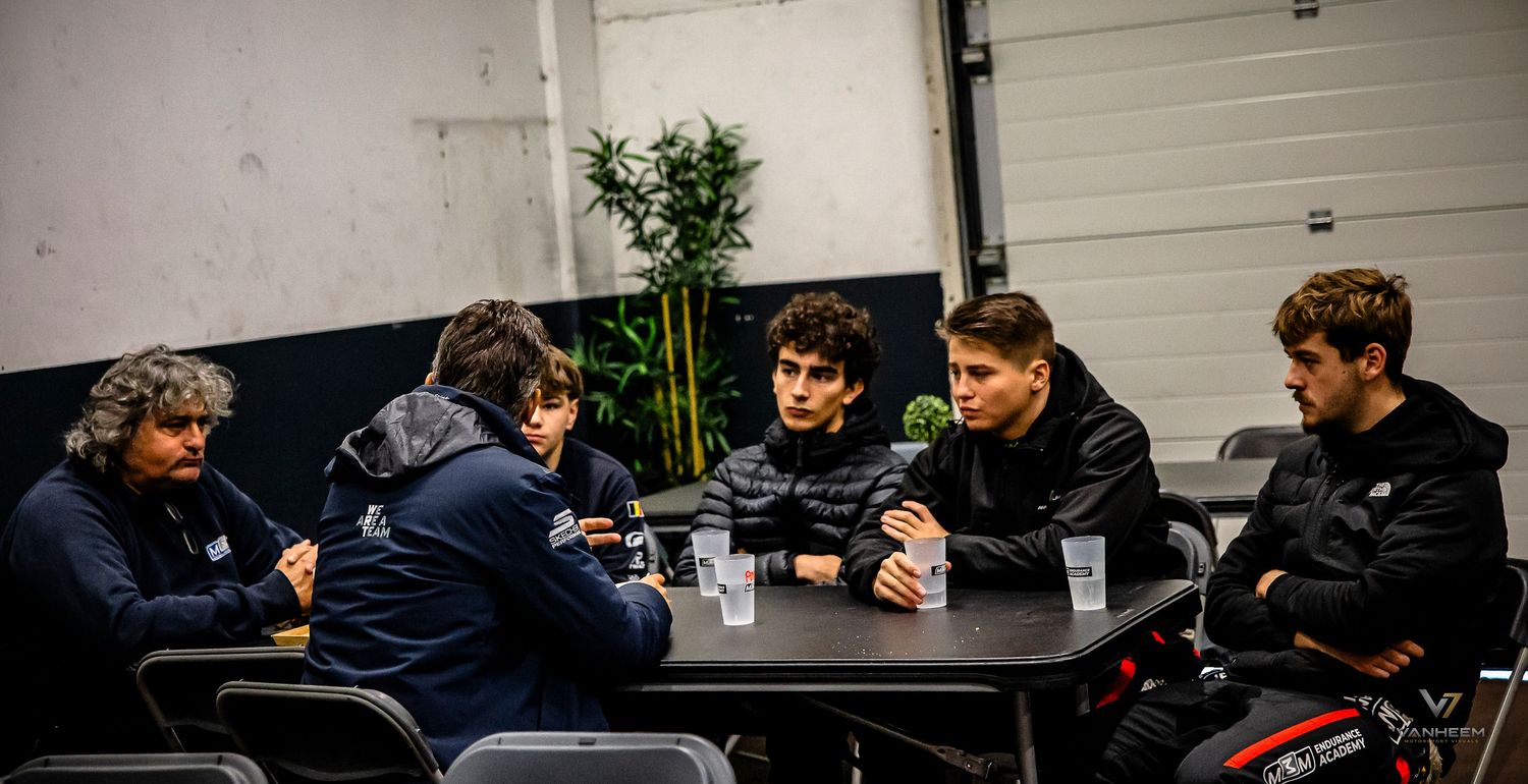 Group of young men sitting around a black table in a garage, engaged in conversation with drinks nearby.