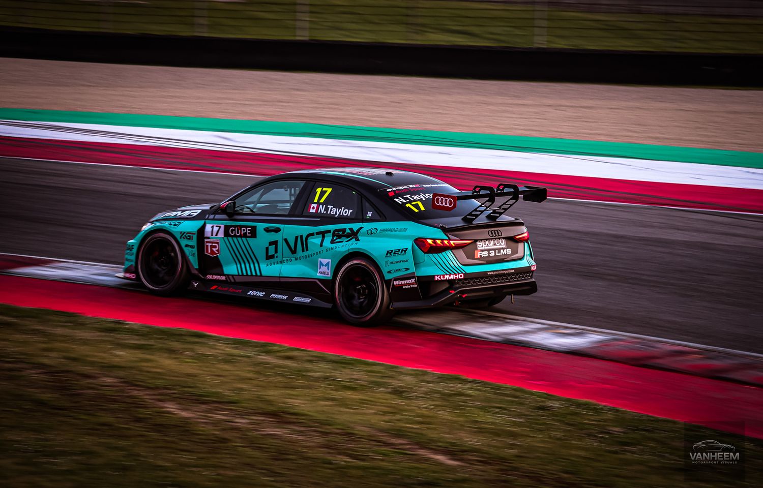 Teal racing car speeds around a racetrack with red and white curbing during a motorsport competition.