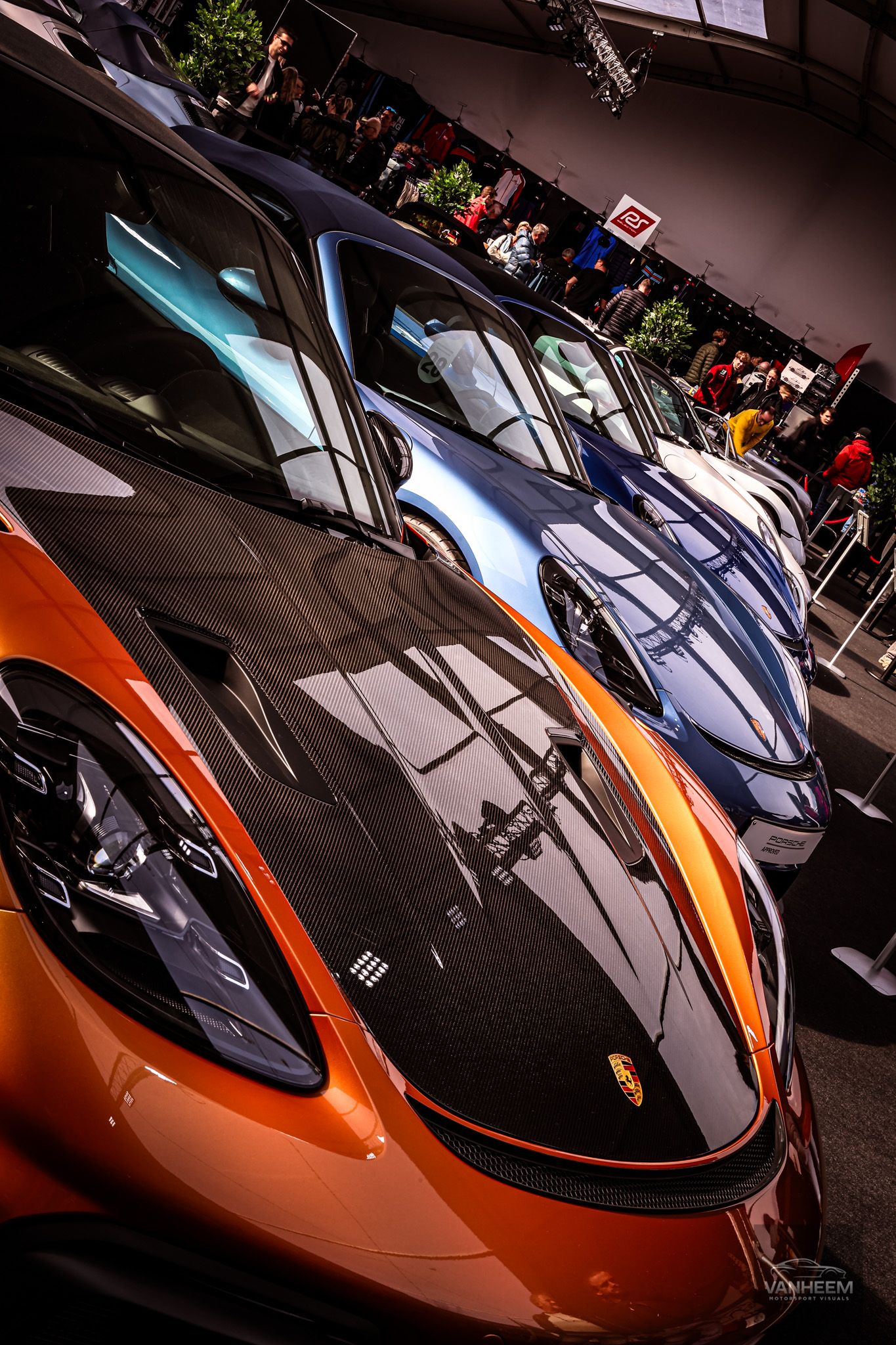 Row of luxury Porsche sports cars on display at an auto show, with a vibrant orange model in the foreground.