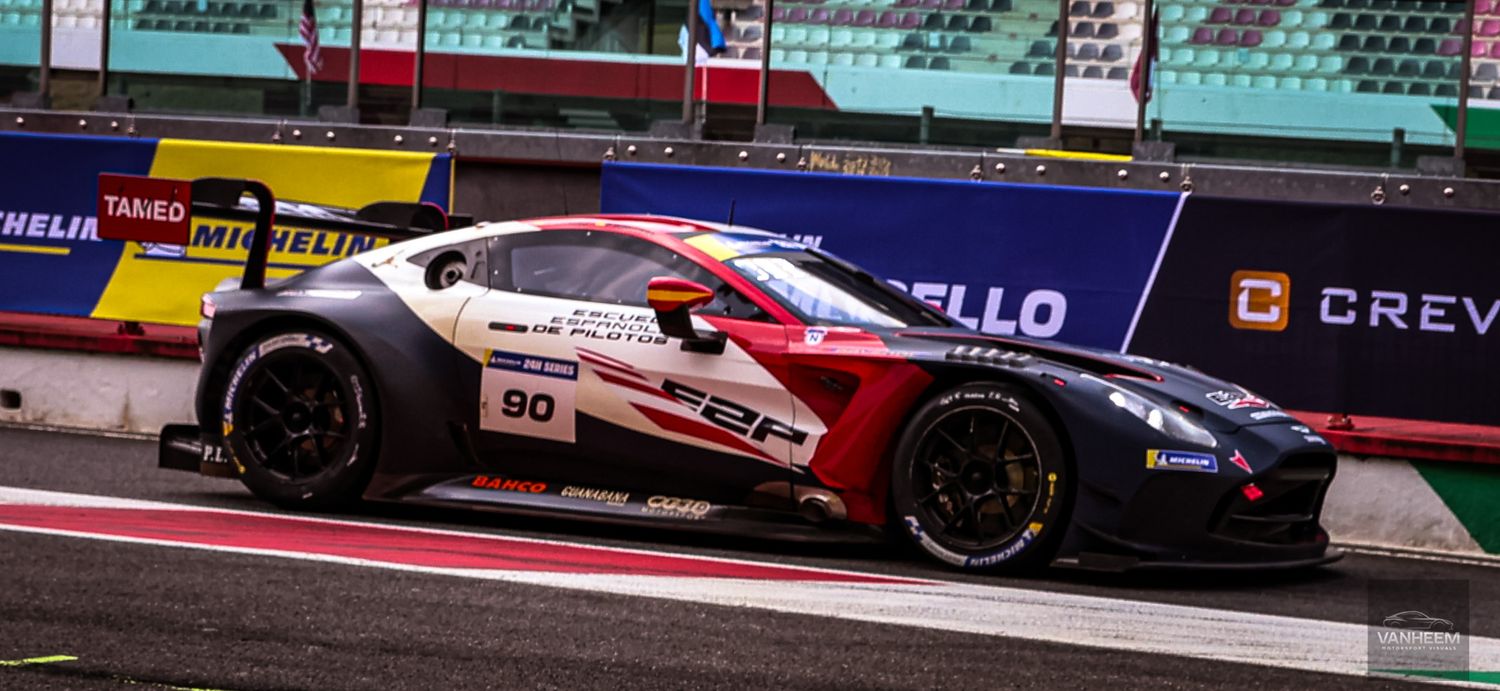 Aston Martin GT3 race car number 90 on track at a motorsport circuit with colorful advertising boards.