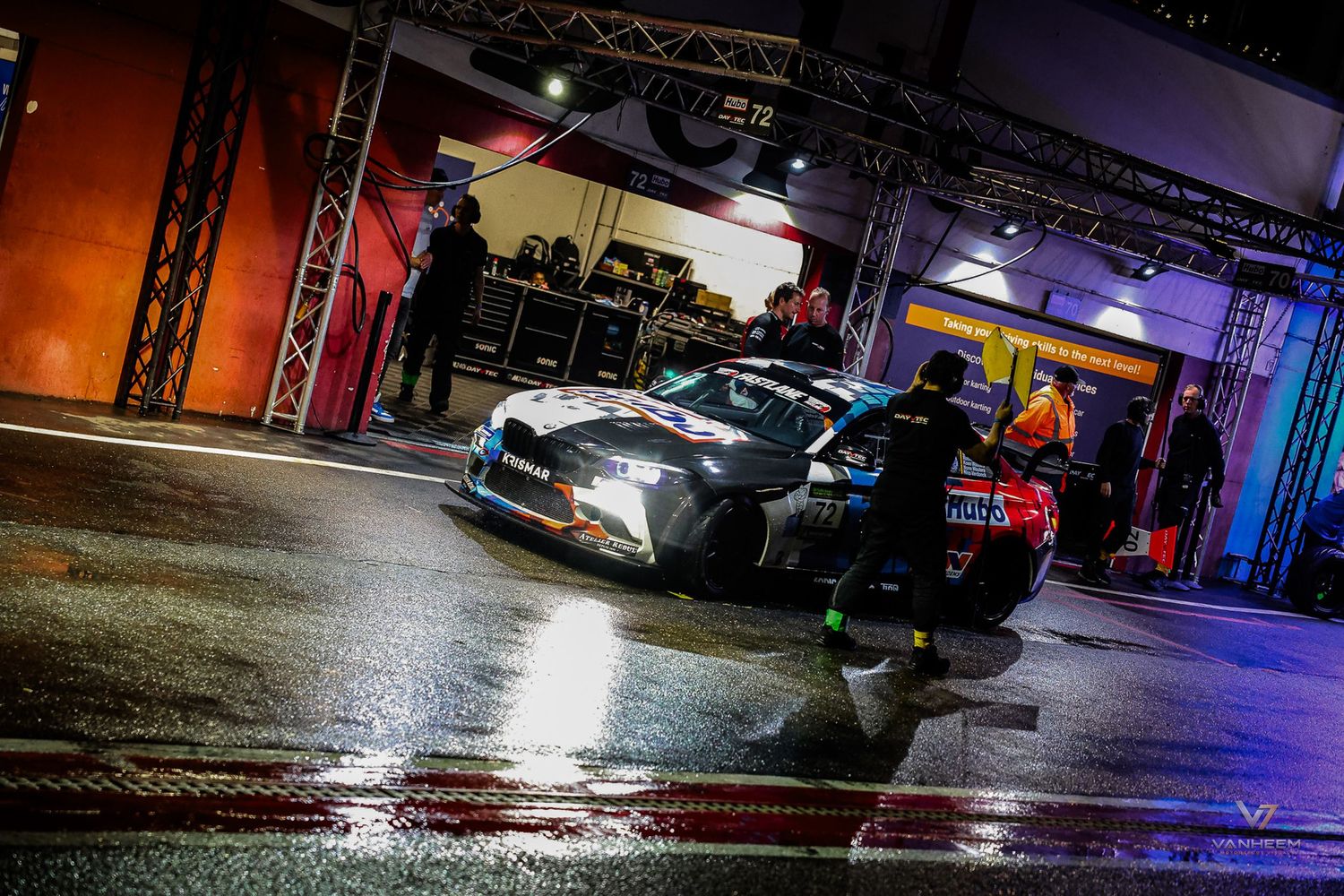 A racing car exits the pit lane at night during a motorsport event, illuminated by bright lights reflecting on wet pavement.