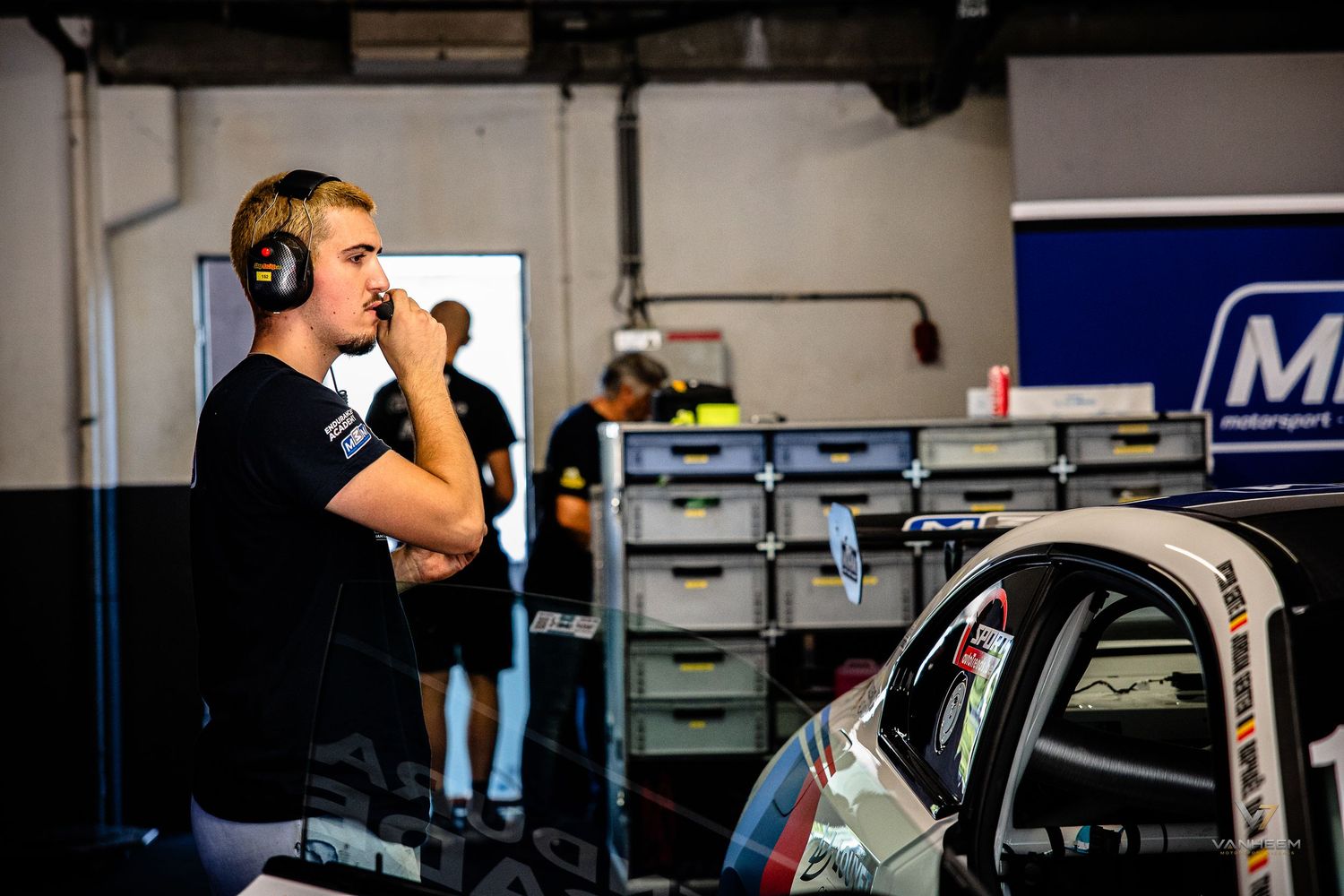 Young racing driver stands in a motorsport garage pit lane, wearing headphones, beside a race car.