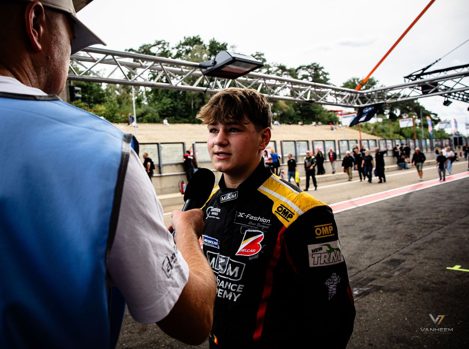 Young racing driver in team uniform being interviewed at a motorsport event paddock area.