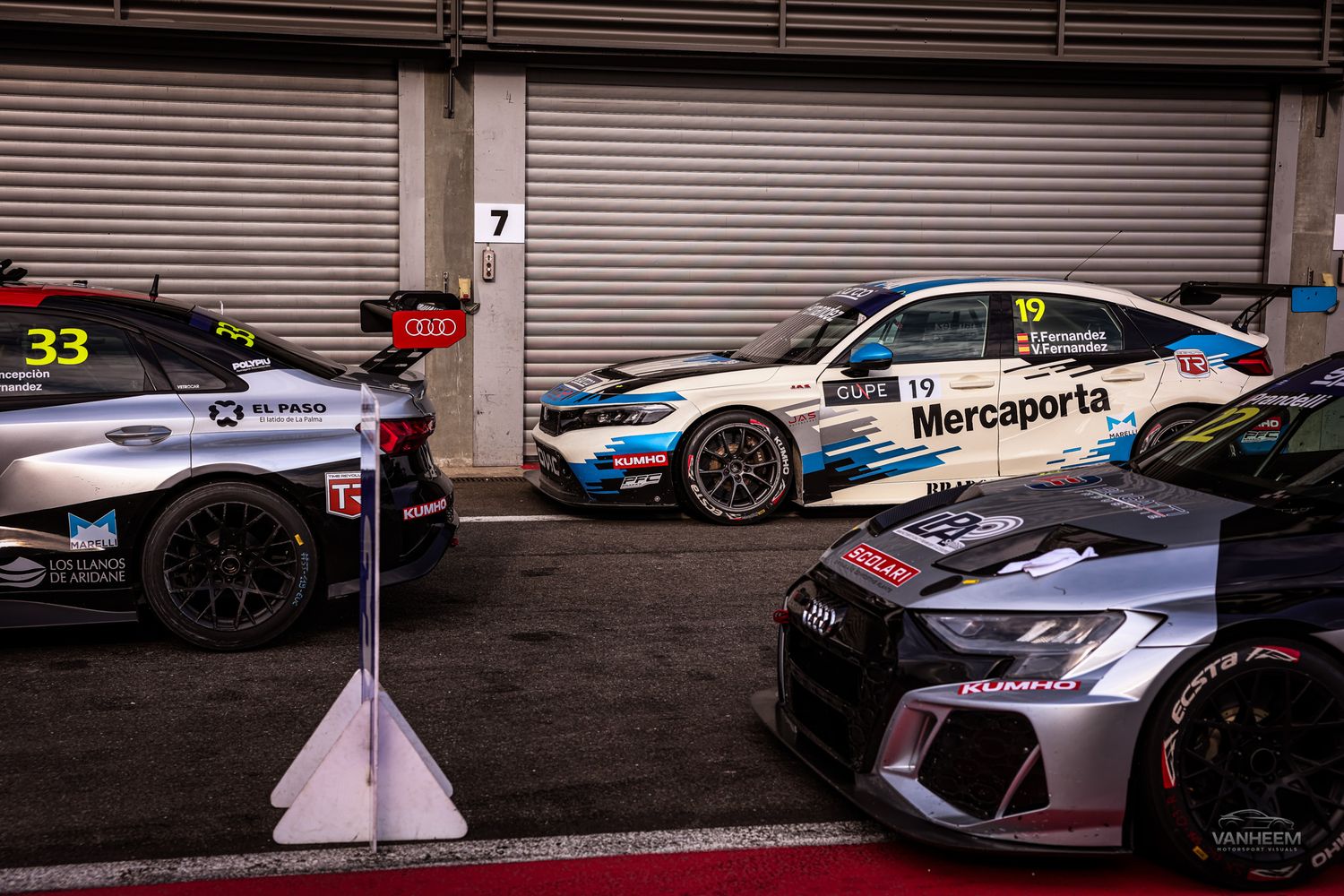 Racing cars lined up in pit lane garage, including Mercaporta #19 and #33 Audi touring cars at a motorsport event.