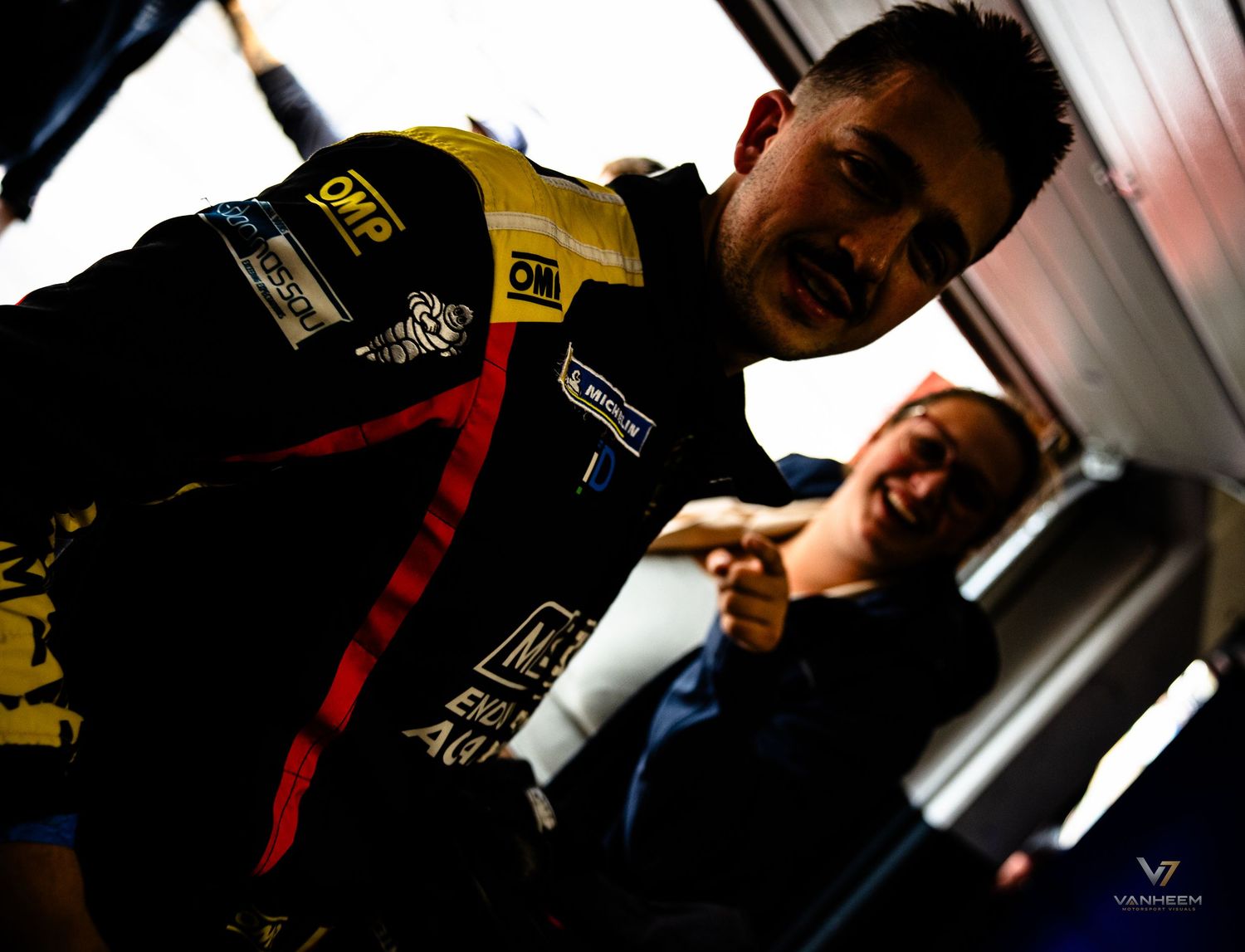 Racing driver in black OMP suit laughing with crew member in dimly lit garage paddock area.