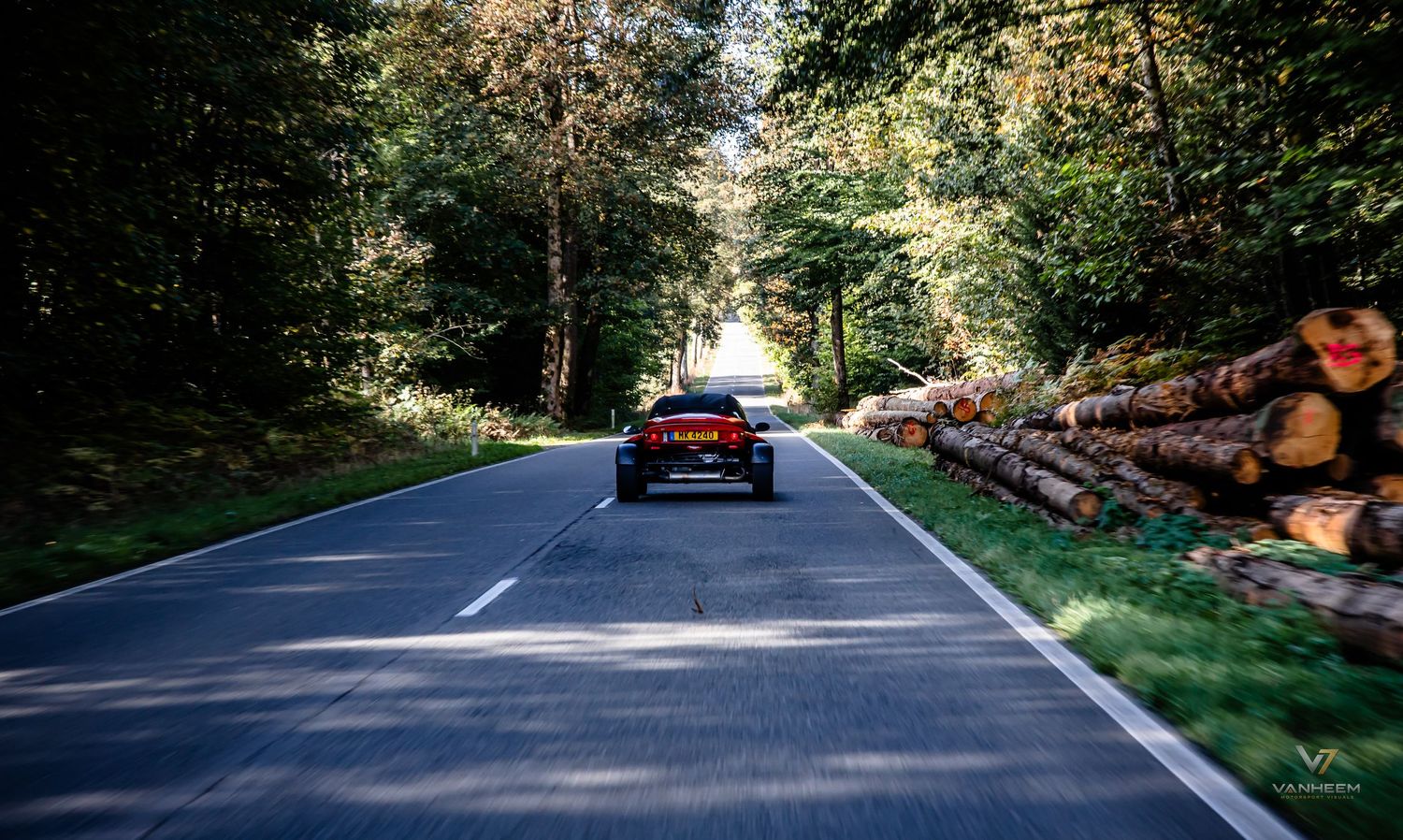 A red sports car drives along a winding forest road lined with stacked logs and lush green trees.