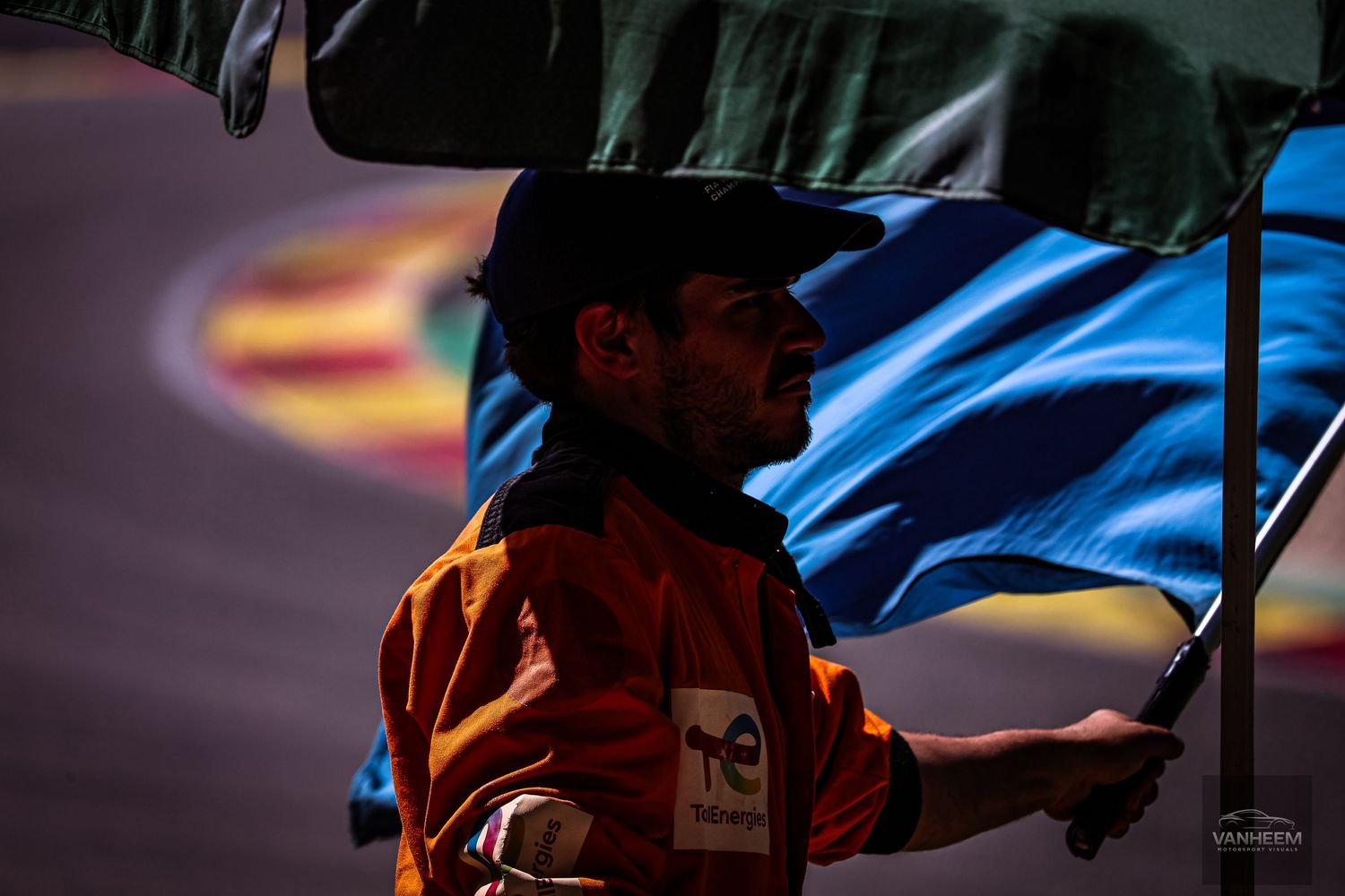 A race marshal in an orange uniform holds a blue flag under an umbrella at a motorsport circuit.