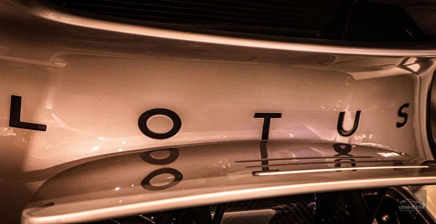 Close-up of Lotus car logo emblem on a sleek silver sports car body panel under warm lighting.