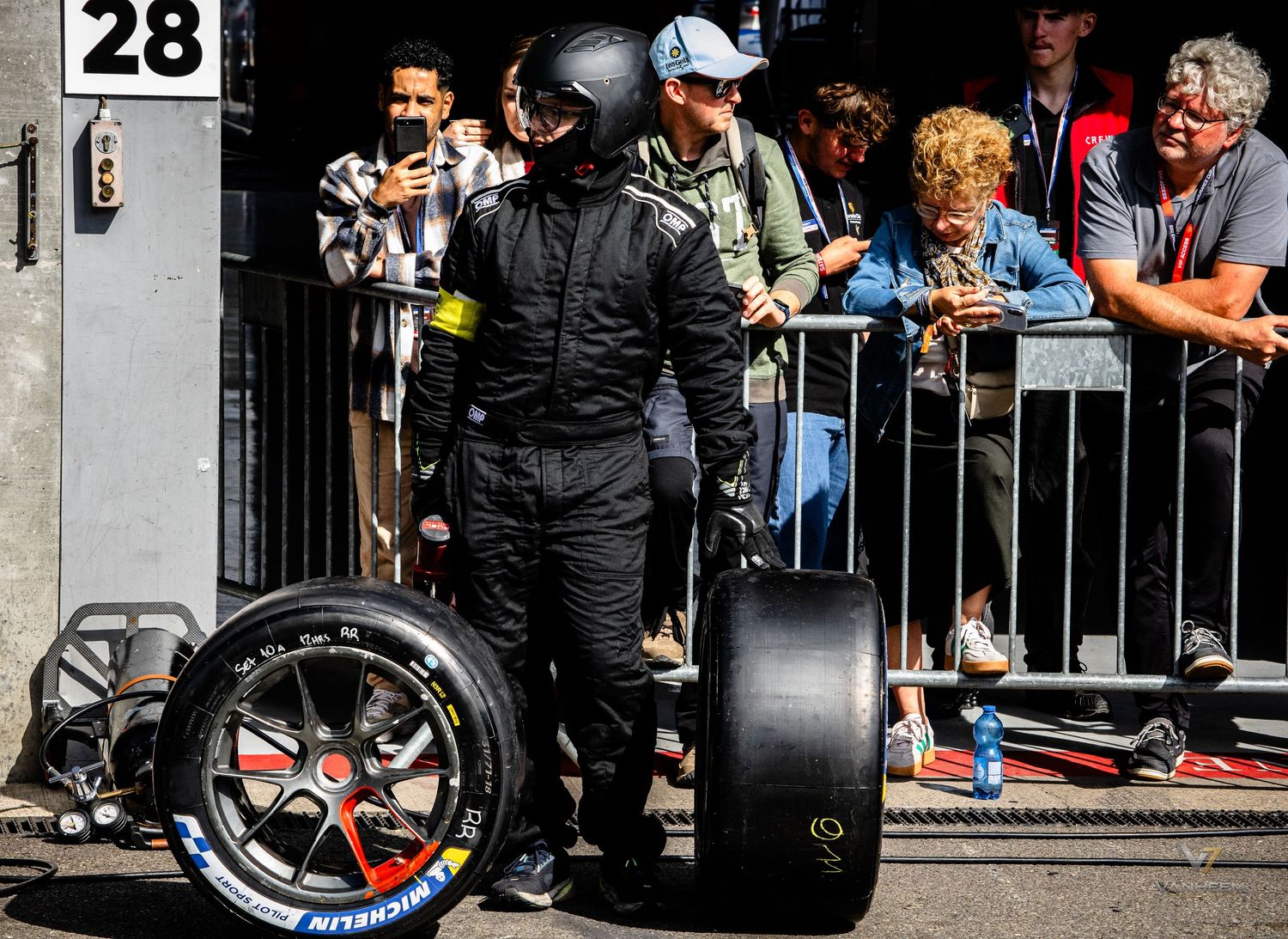 A pit crew member in black racing suit handles Michelin tires in the pit lane while spectators watch from behind barriers.