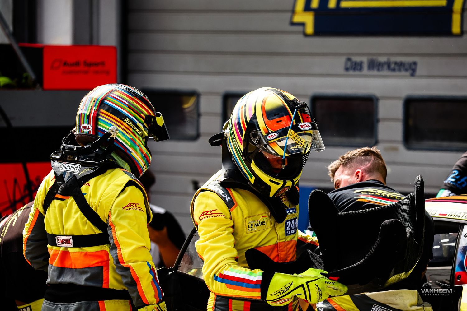 Two racing drivers in yellow helmets and suits stand in pit lane during a motorsport event.