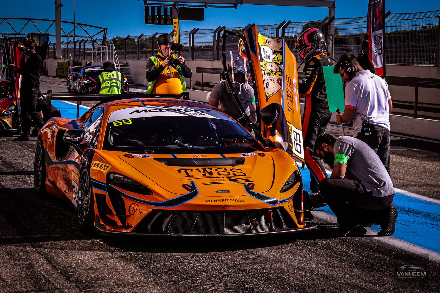 Orange McLaren GT3 race car in pit lane with mechanics performing service during a motorsport racing event.