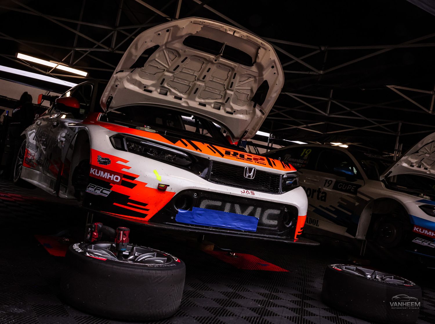 Honda Civic racing car with hood open undergoing maintenance in a motorsport garage pit area.