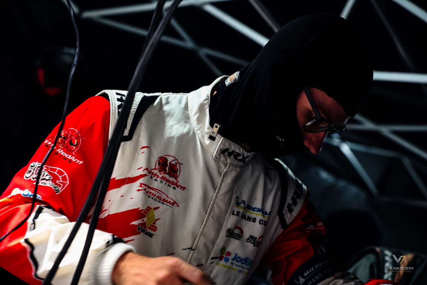 Race car driver in white and red suit with balaclava working in pit garage under dramatic lighting.