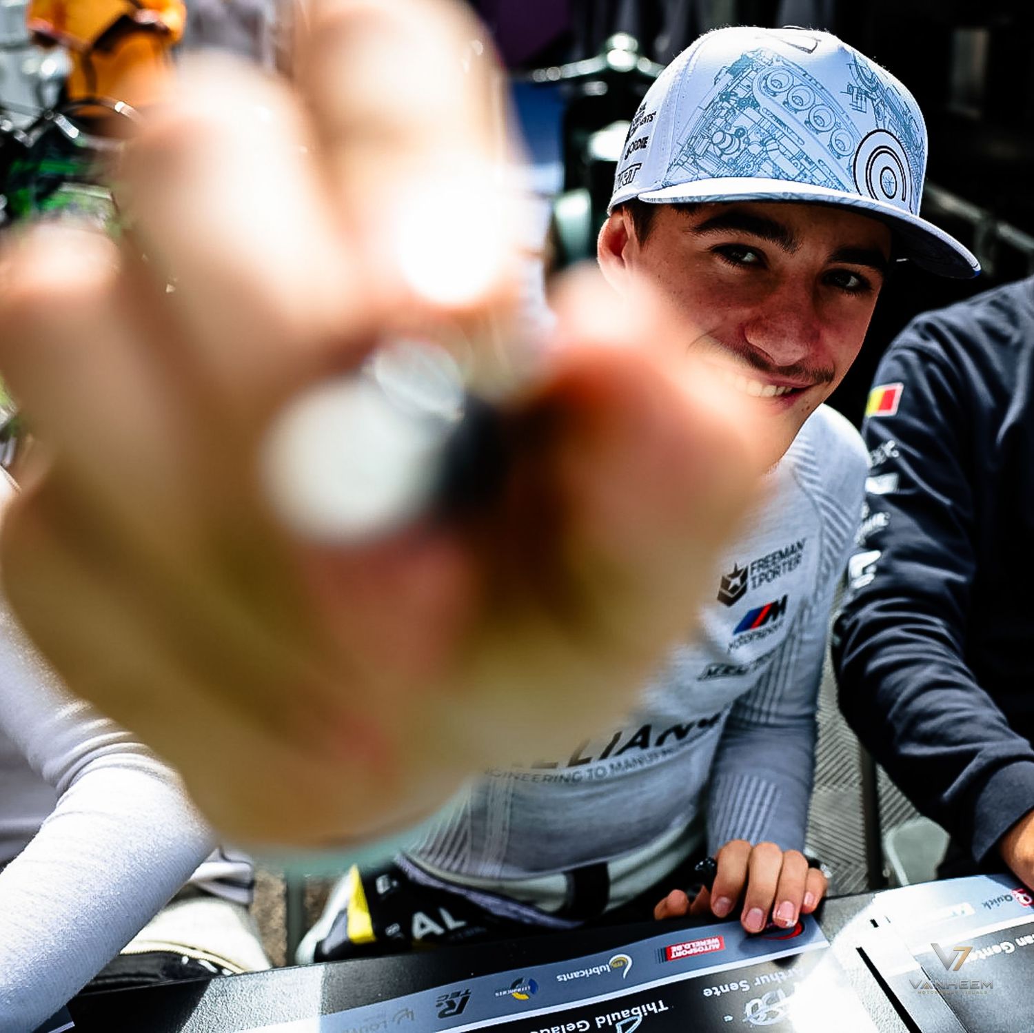 Young racing driver in cap smiles while holding coin toward camera at motorsport event pit lane.
