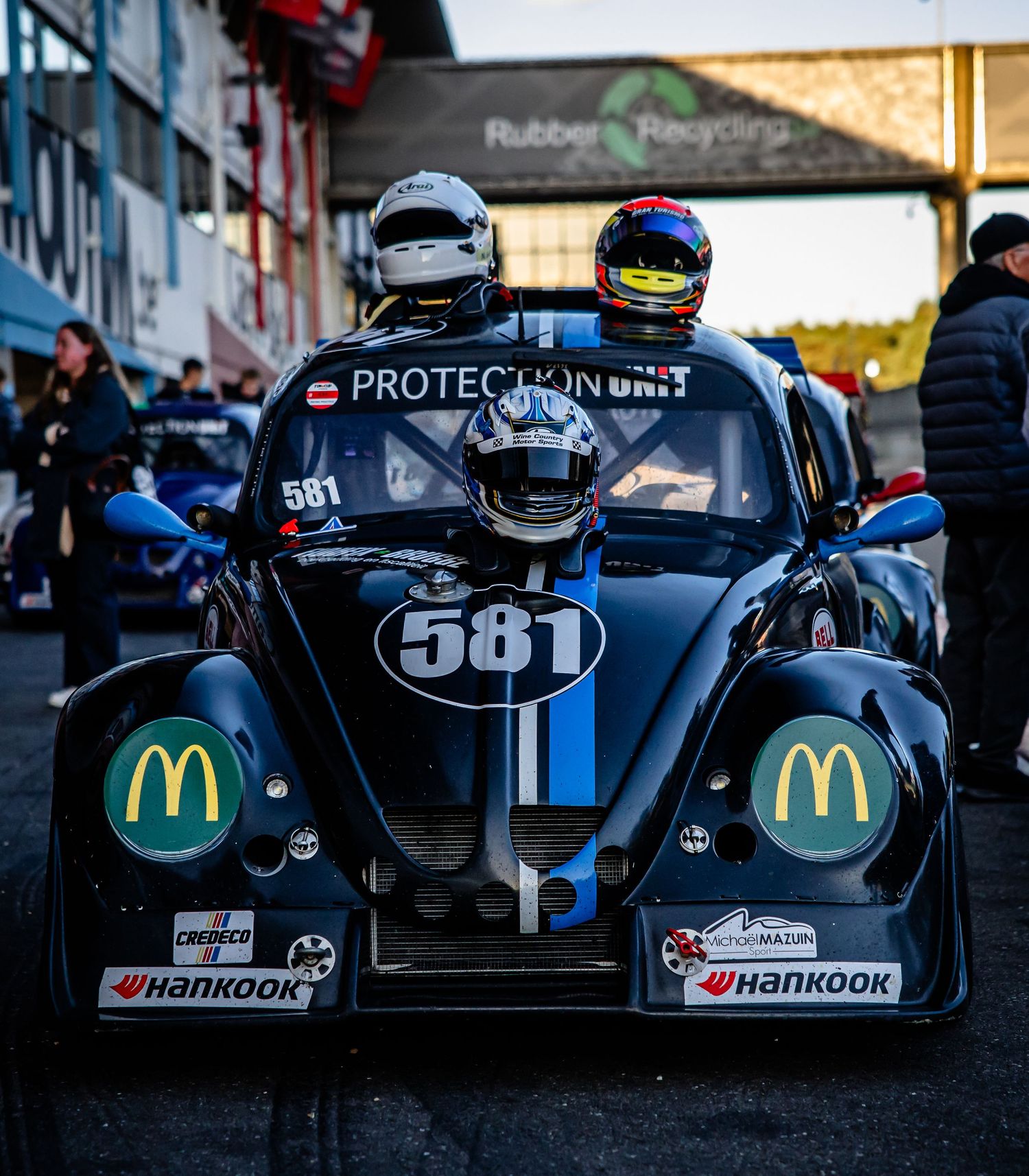 Two helmeted drivers sit in black race car 581 with McDonald's and Hankook sponsors in pit lane.
