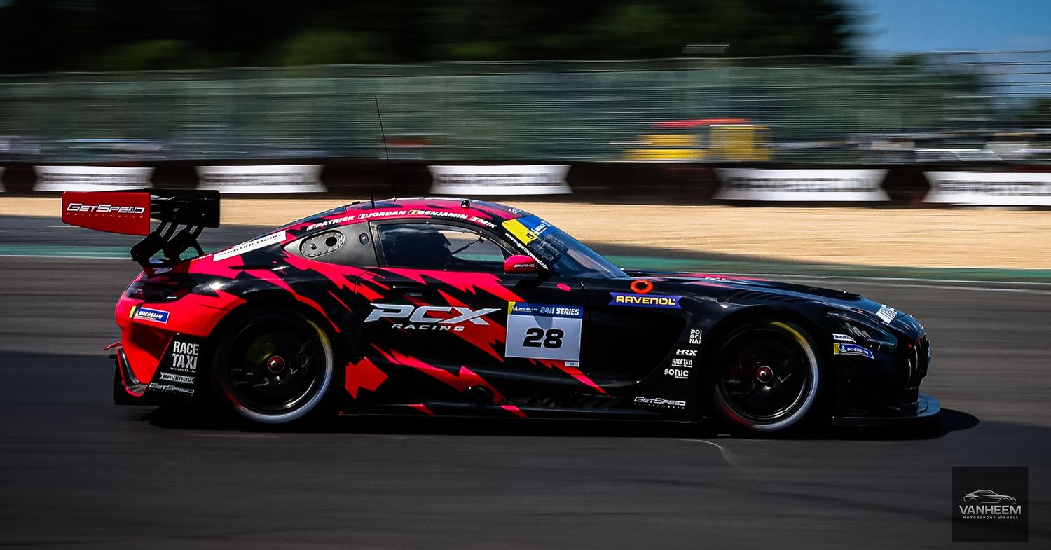 Red and black GT race car number 28 competing on track during a sunny motorsport event.