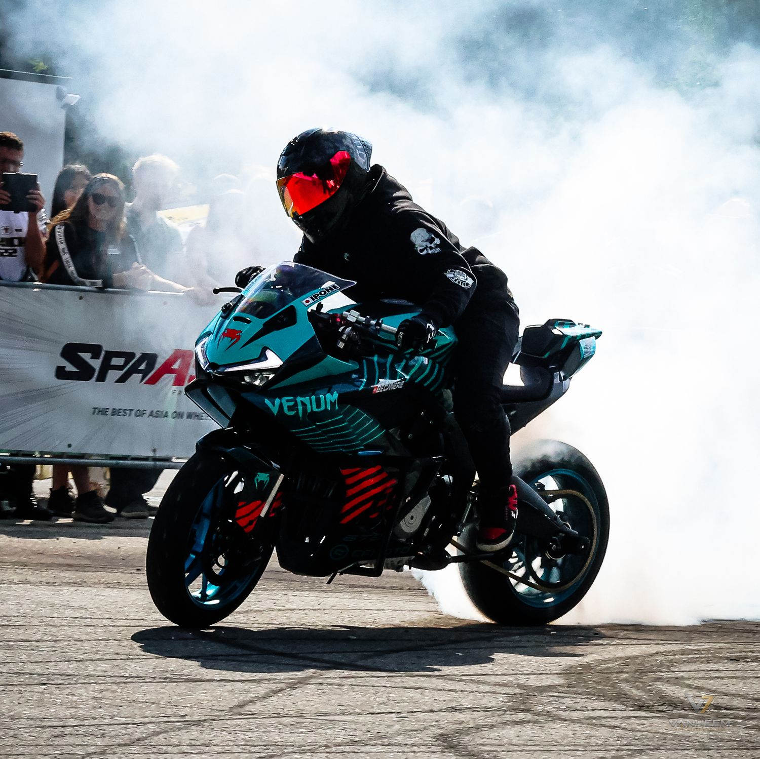 Motorcyclist performs a smoky burnout on a teal Venom sport bike at a racing event with spectators watching.