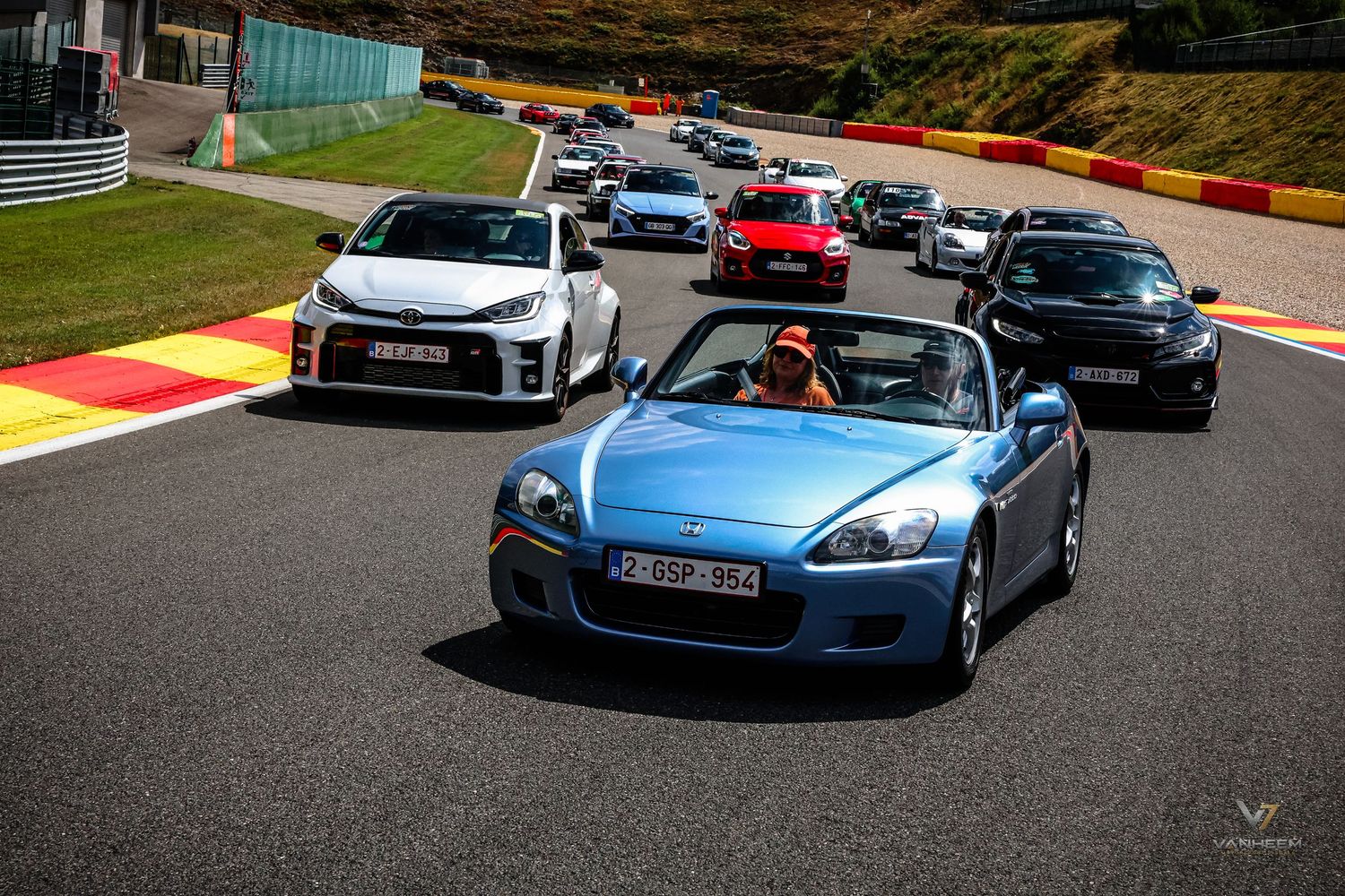 A blue Honda S2000 leads a convoy of sports cars on a racing circuit track on a sunny day.
