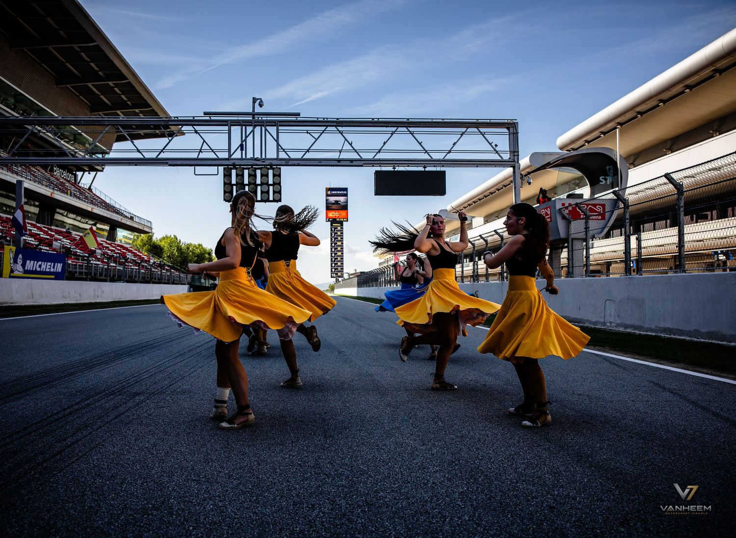 Dancers in yellow skirts perform on a motorsport circuit pit lane at dusk with Michelin signage in the background.