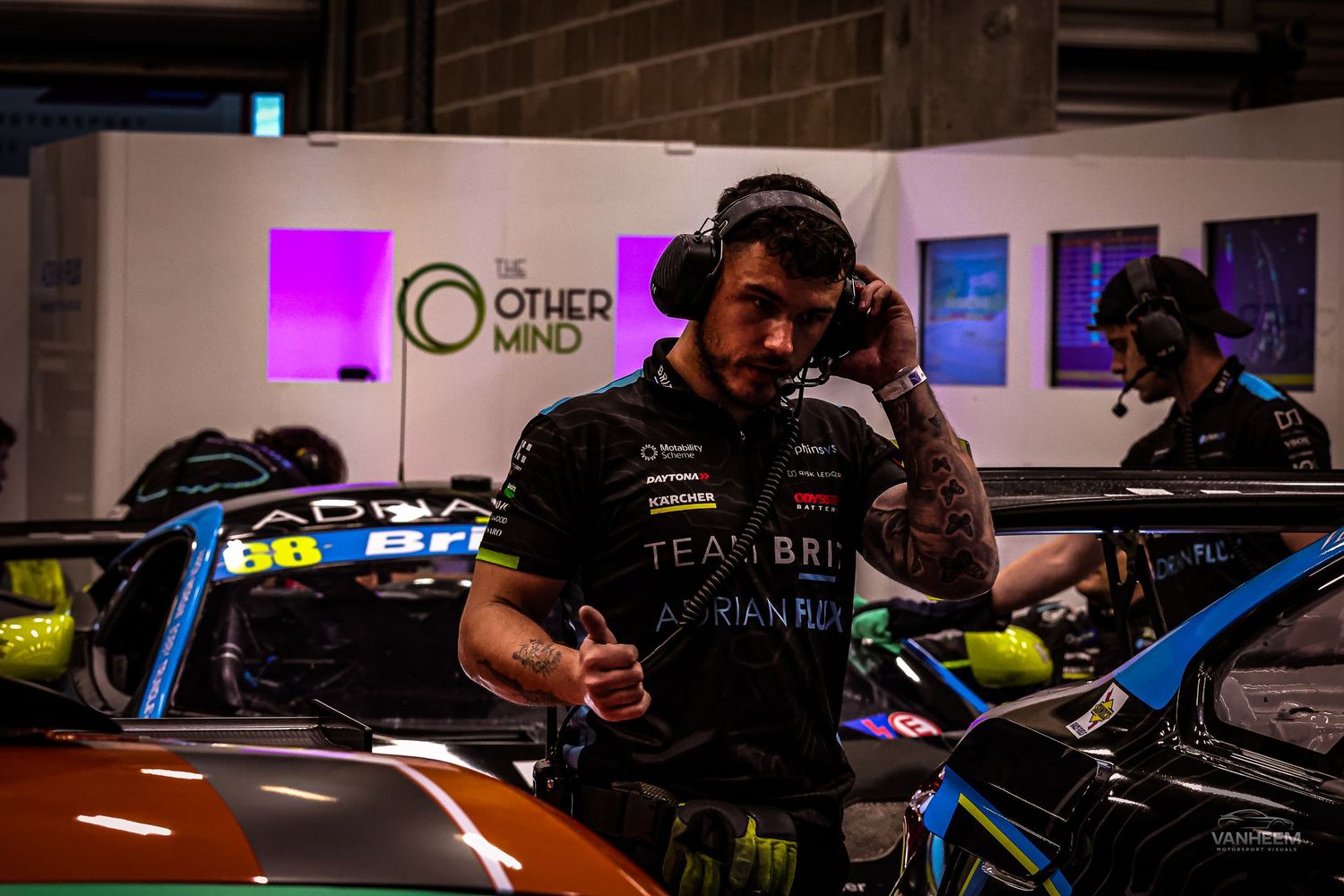 A Team Brit racing driver in the pit garage wearing headphones and team gear, giving a thumbs up near race cars.
