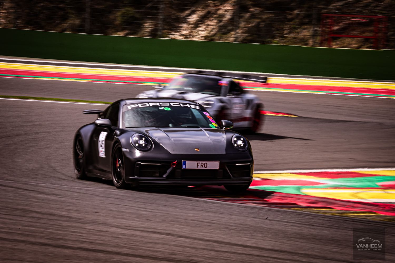 A dark Porsche 911 GT3 races on a circuit track with colorful rumble strips visible in the background.