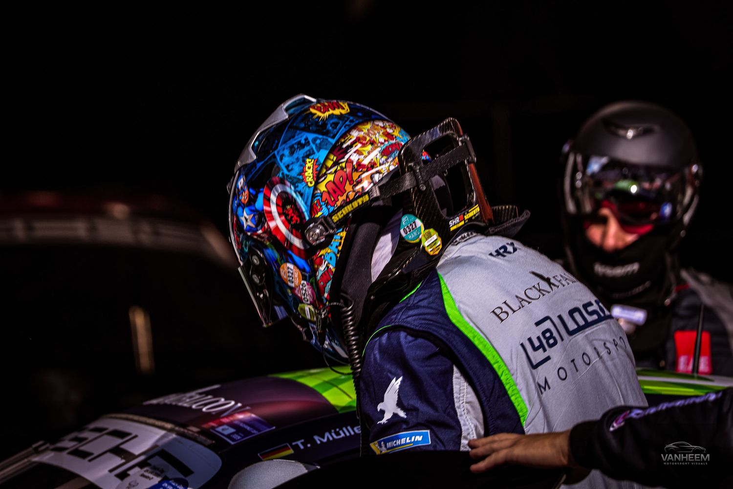 Racing driver wearing a colorful graffiti-style helmet and team racing suit at a nighttime motorsport event.