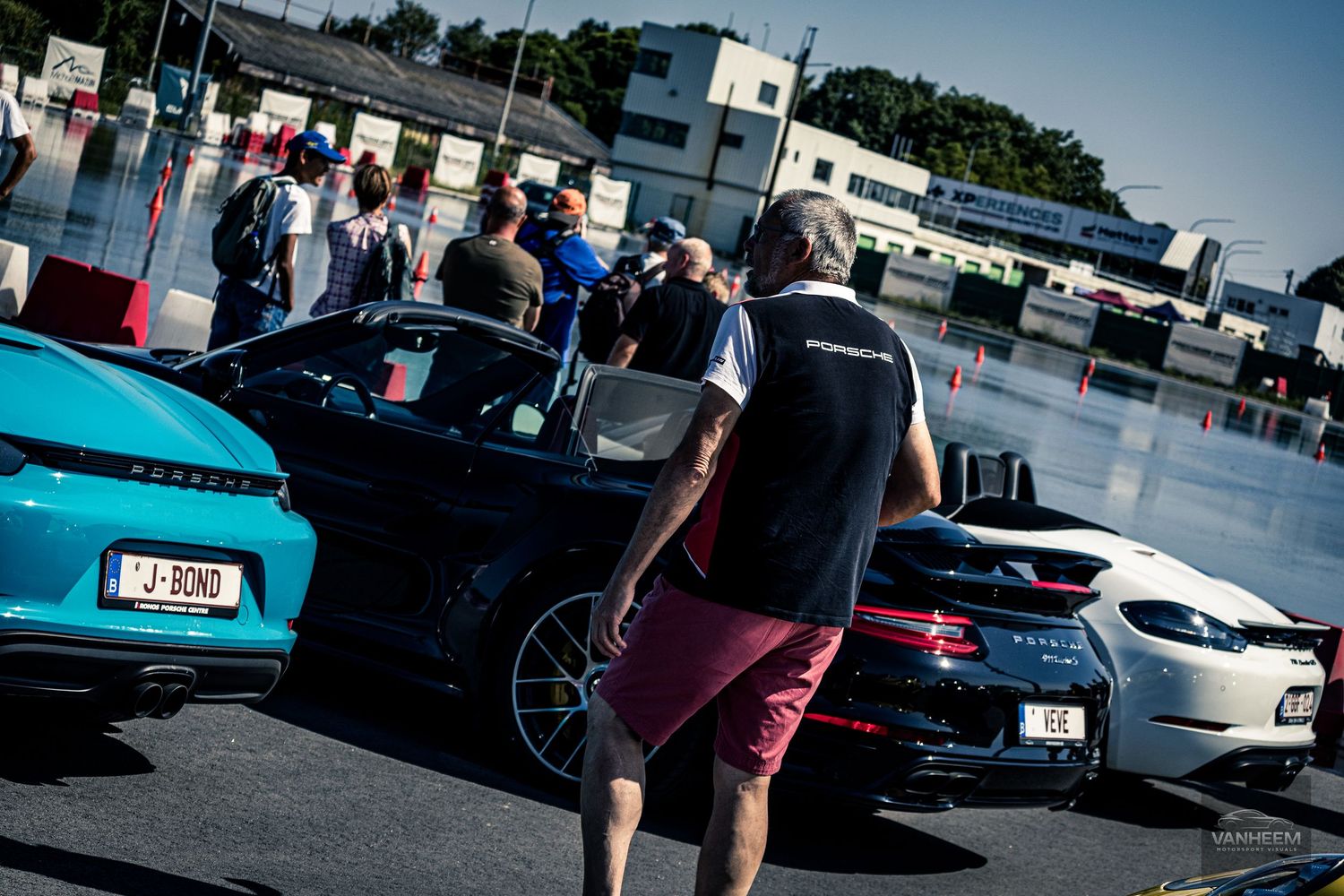 Cars lined up at an outdoor automotive event with people walking between vehicles on a sunny day.