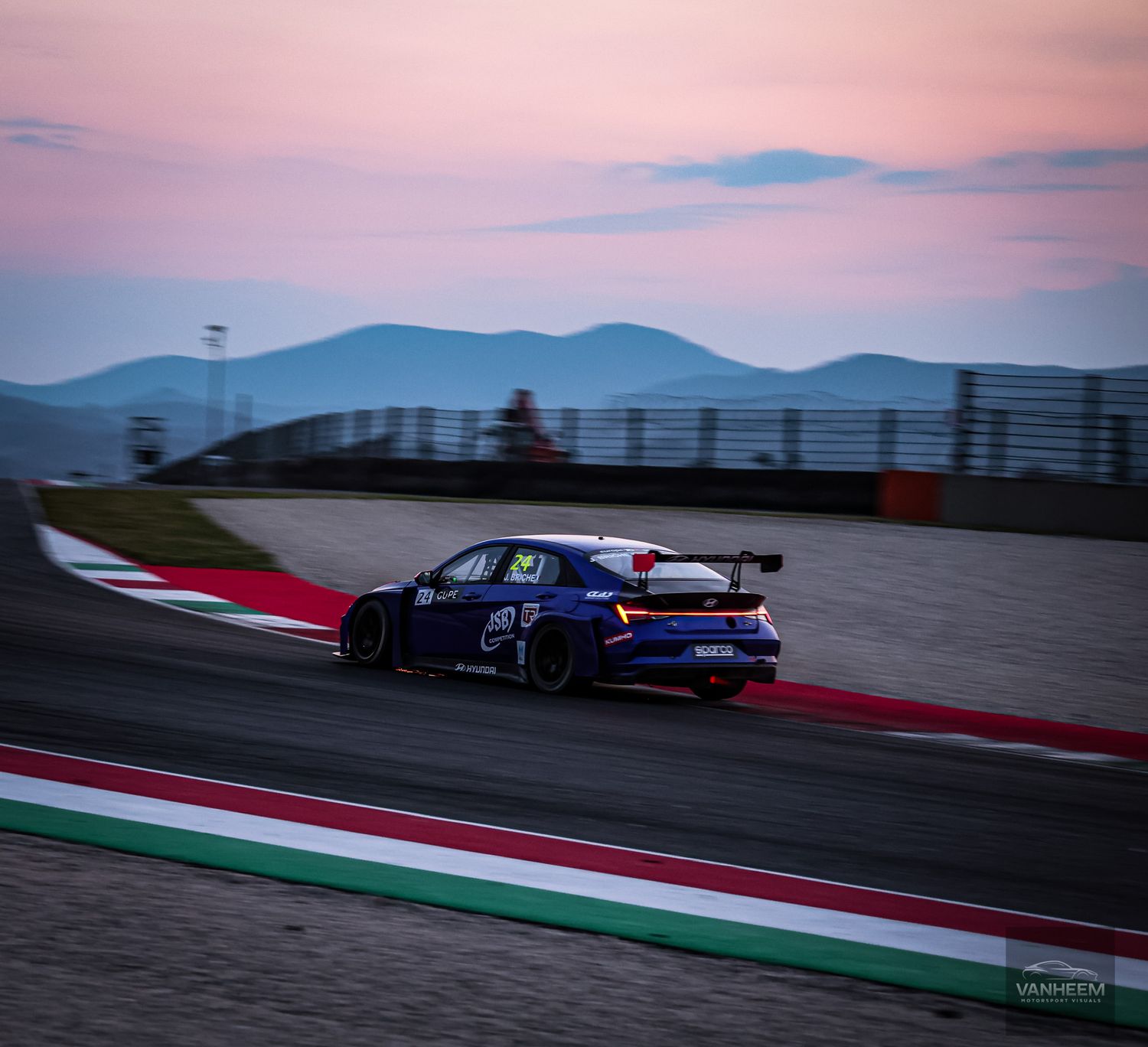 Race cars speed through a winding track during sunset with mountains in the background.