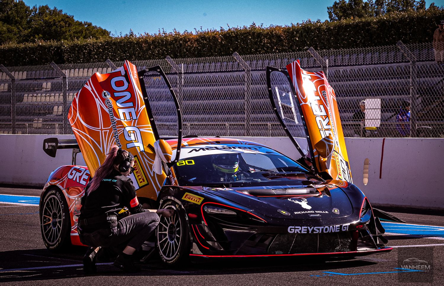 Racing cars line up on a track during sunset with colorful team flags and banners visible in the background.