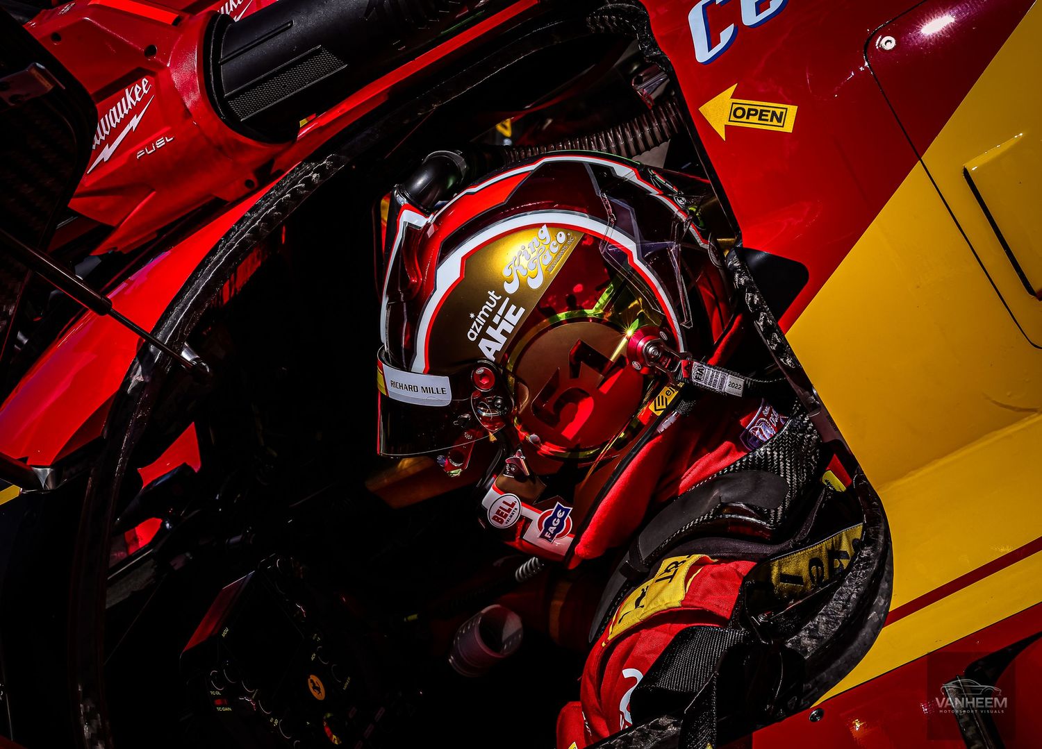 Race car driver in red helmet and suit seated inside a vibrant red and yellow racing vehicle cockpit.