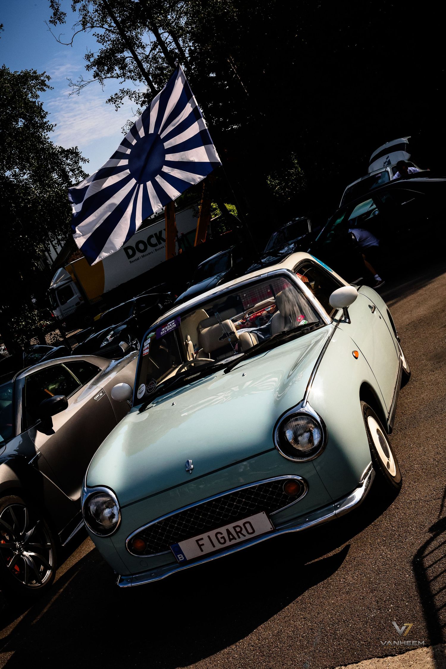 A mint green Nissan Figaro classic car displayed at an outdoor car show with a rising sun flag in the background.