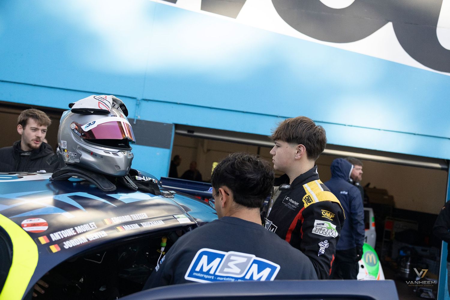 Racing drivers in pit lane discuss strategy, one holding a helmet, wearing branded race suits near a competition vehicle.