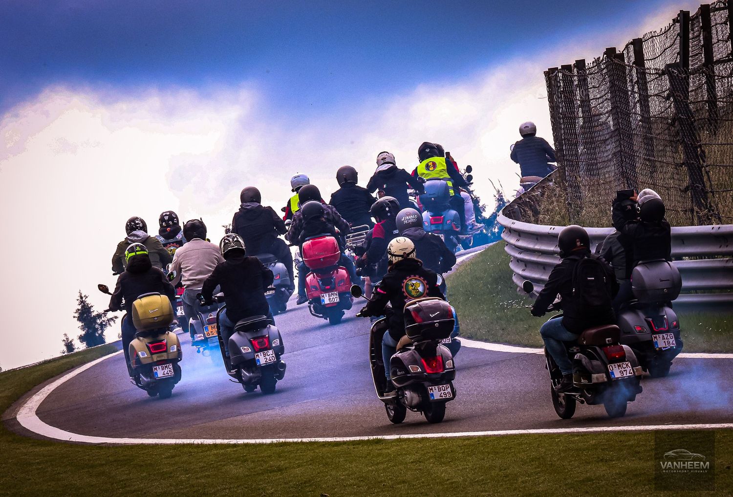 Group of motorcyclists riding scooters on a winding racetrack curve under dramatic cloudy skies.