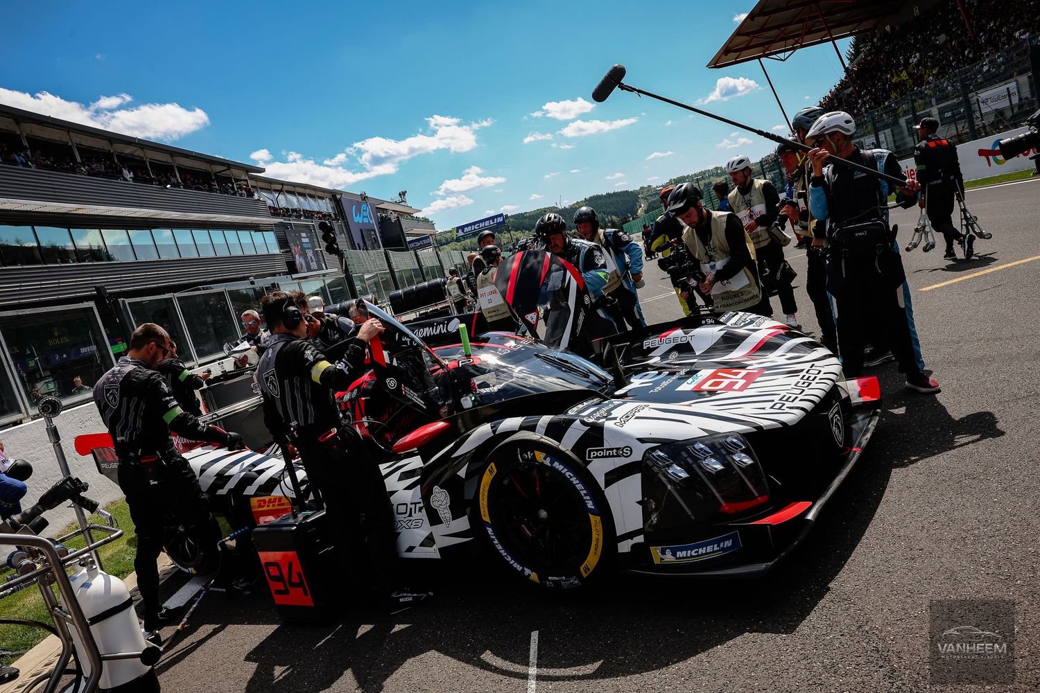 Porsche LMP race car in pit lane surrounded by mechanics during a motorsport event on a sunny day.