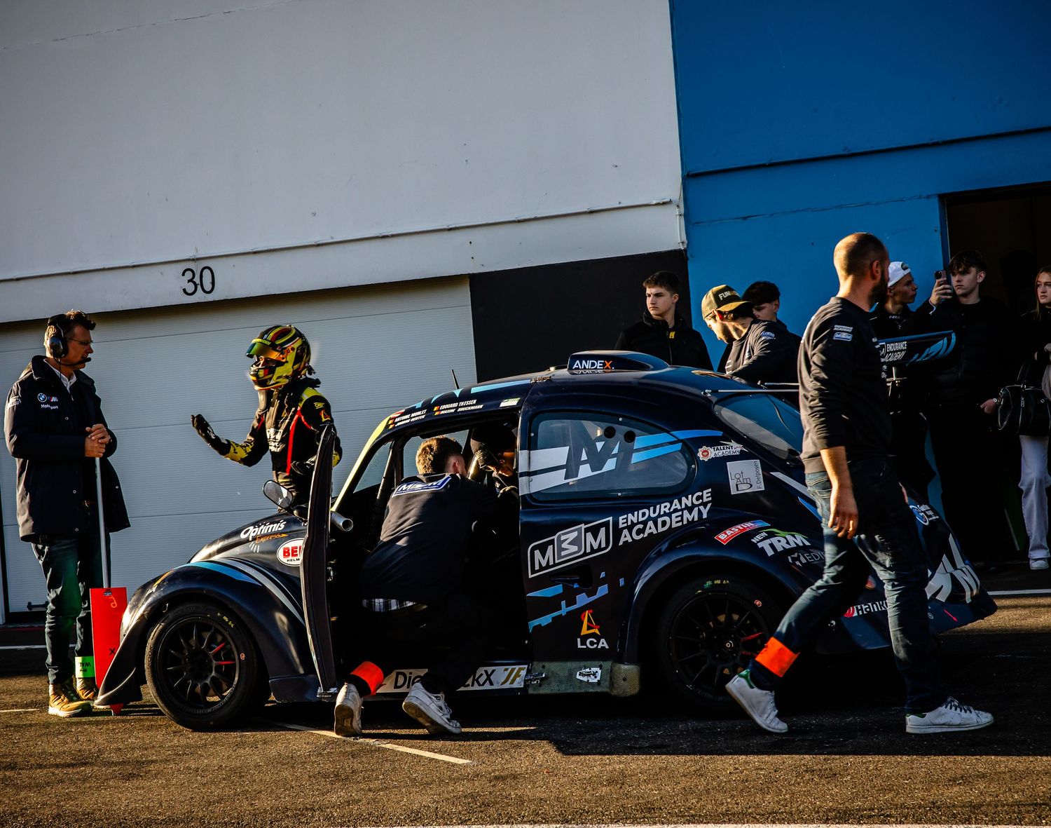 Racing team crew members surround a black race car in pit lane during a motorsport event at a circuit.