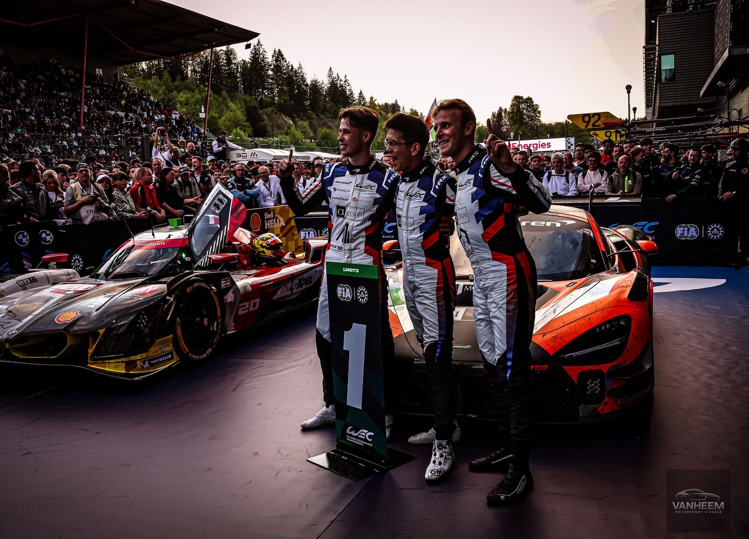 Three racing drivers pose together in front of race cars at a motorsport event, wearing team uniforms with national flags.