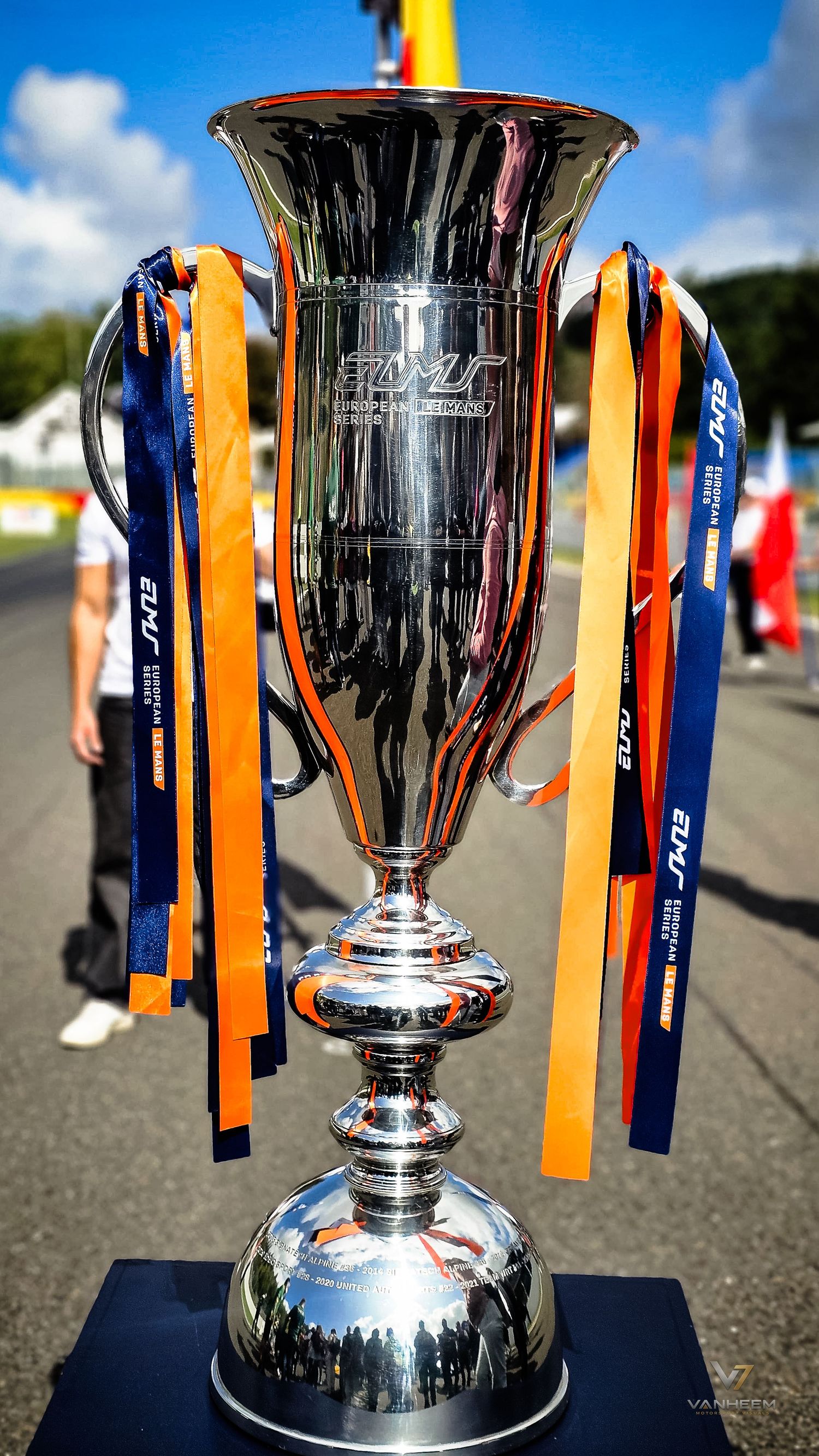 Shiny silver racing trophy with orange and blue ribbons displayed outdoors at a motorsport event on a sunny day.