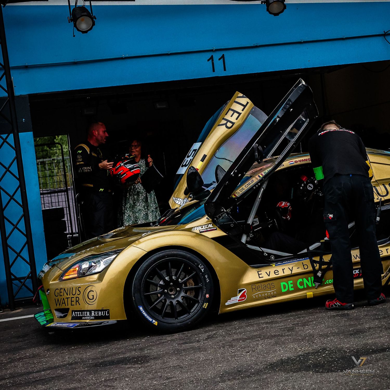 Gold racing car with open doors being serviced by mechanics in pit lane at motorsport event.