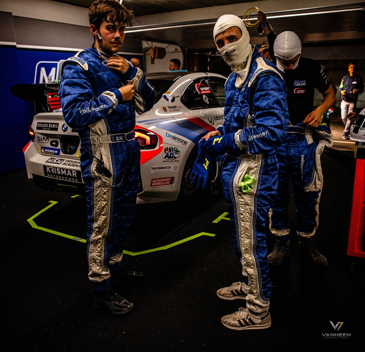 Two racing drivers in blue fire suits stand beside a white BMW race car in a pit garage.