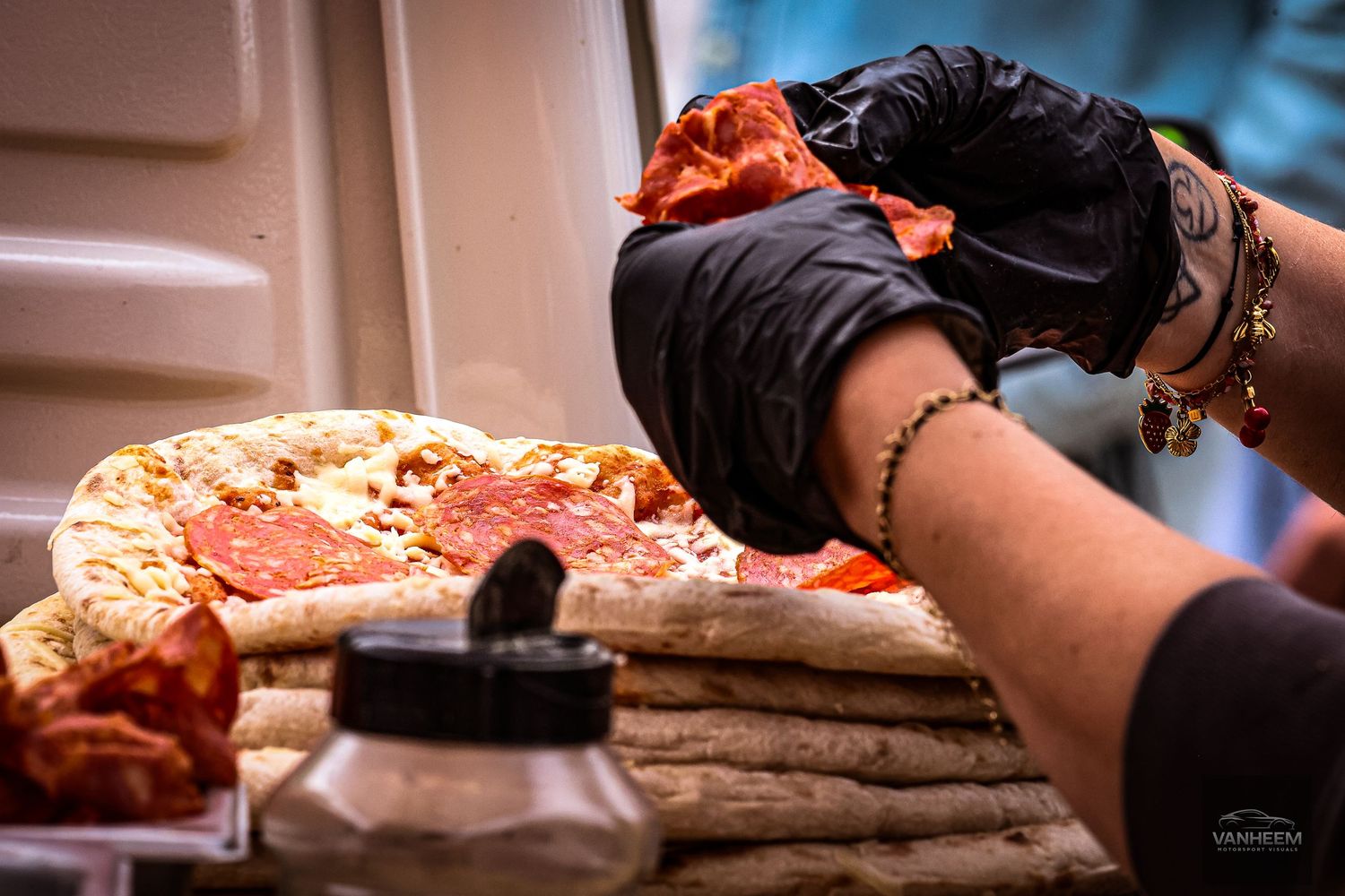 Person in black gloves placing pepperoni on a fresh homemade pizza on a wooden surface.